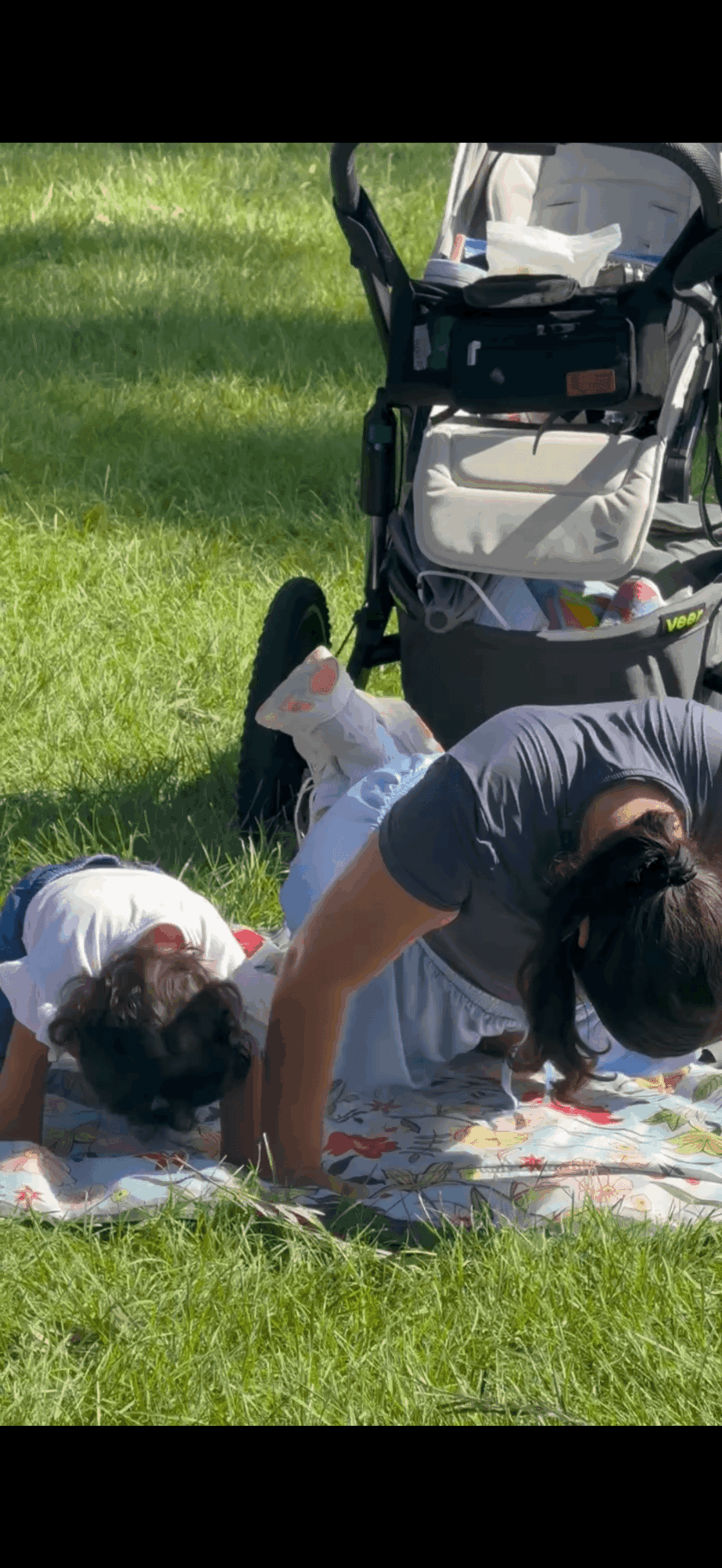 Two children lying on a blanket on grass, with a stroller nearby, outdoors on a sunny day.