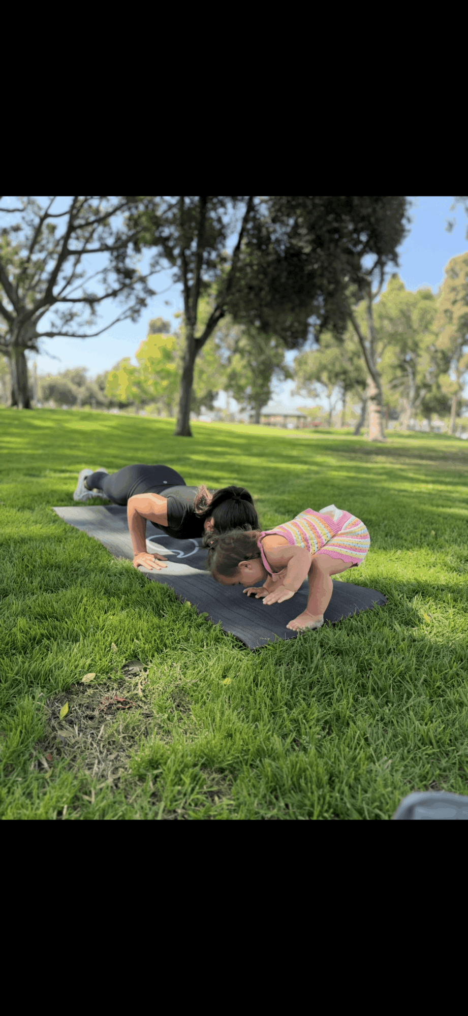 Two children practicing yoga outdoors on a grassy field with trees and blue sky in background.