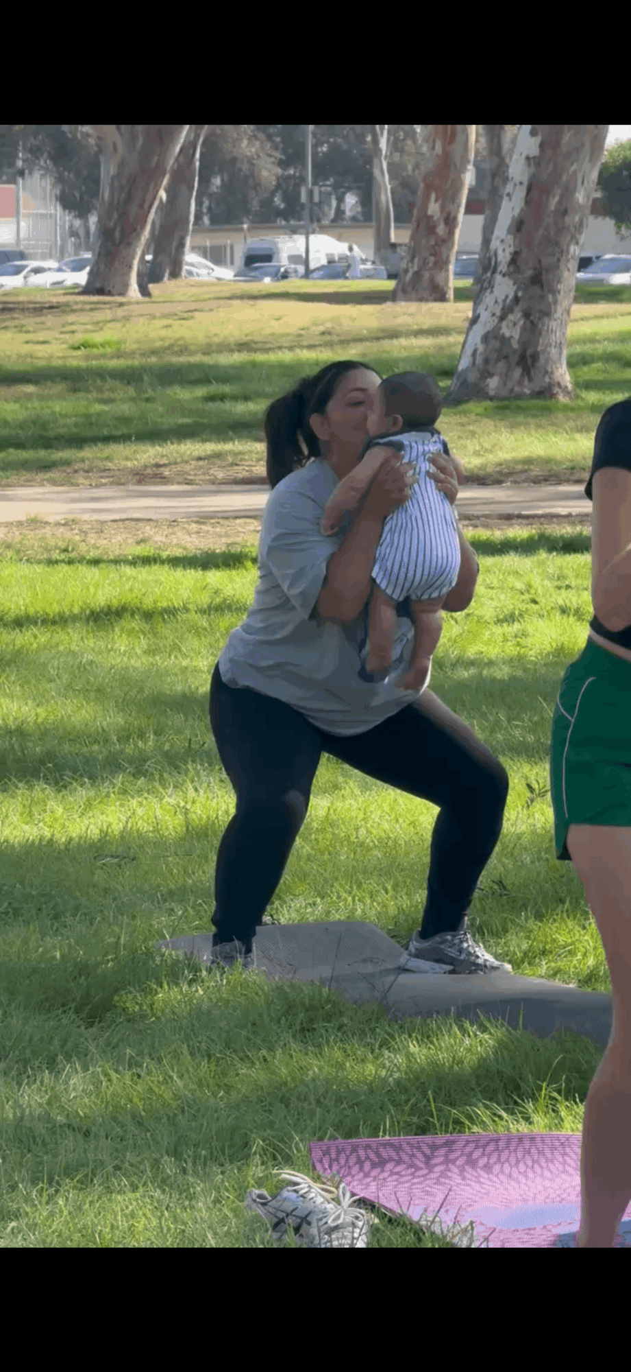 Person holding a baby outdoors in a park with trees and grass, other people partially visible nearby.