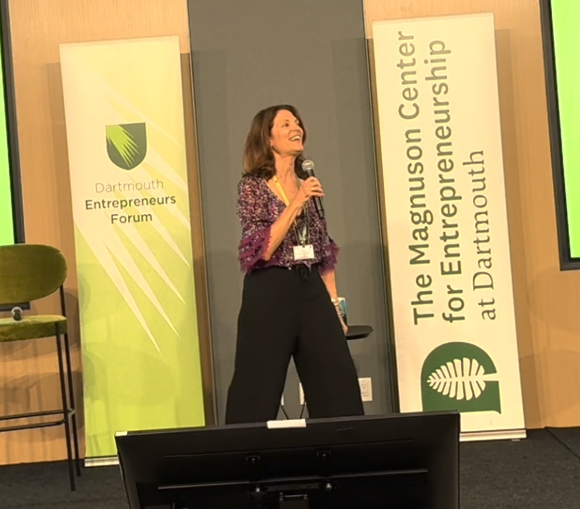 Woman speaking on stage at event, holding microphone, with banners in background, one reading 'Dartmouth Entrepreneurs Forum'.