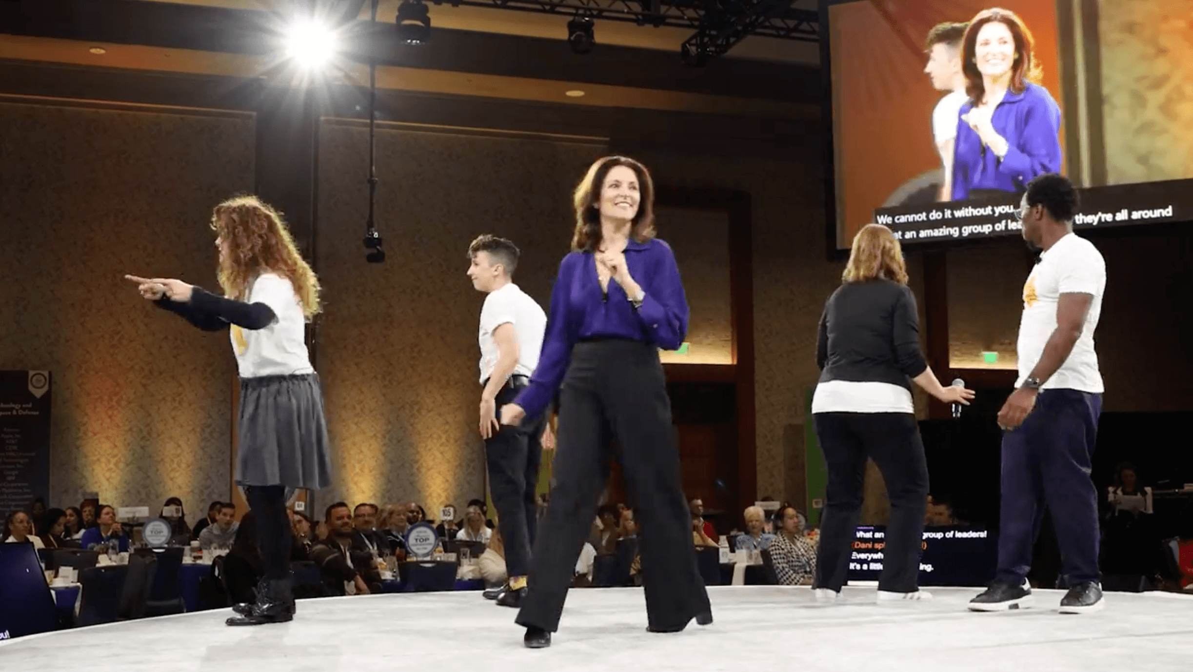 Five people on stage, two women shaking hands, one woman speaking, audience in background, large screen displaying a woman, indoor event