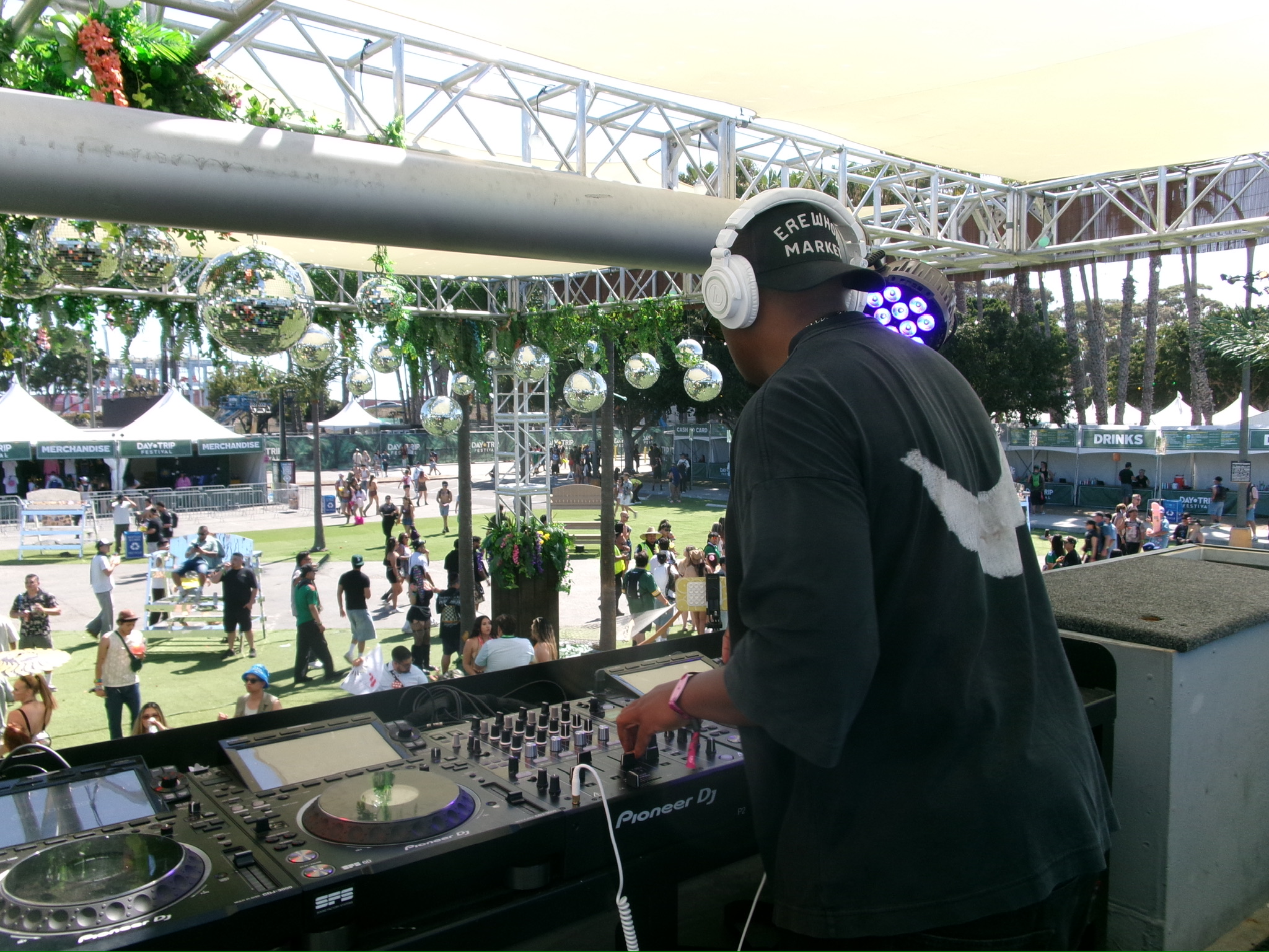 DJ wearing headphones and a cap, operating equipment at an outdoor event with people and tents in the background.
