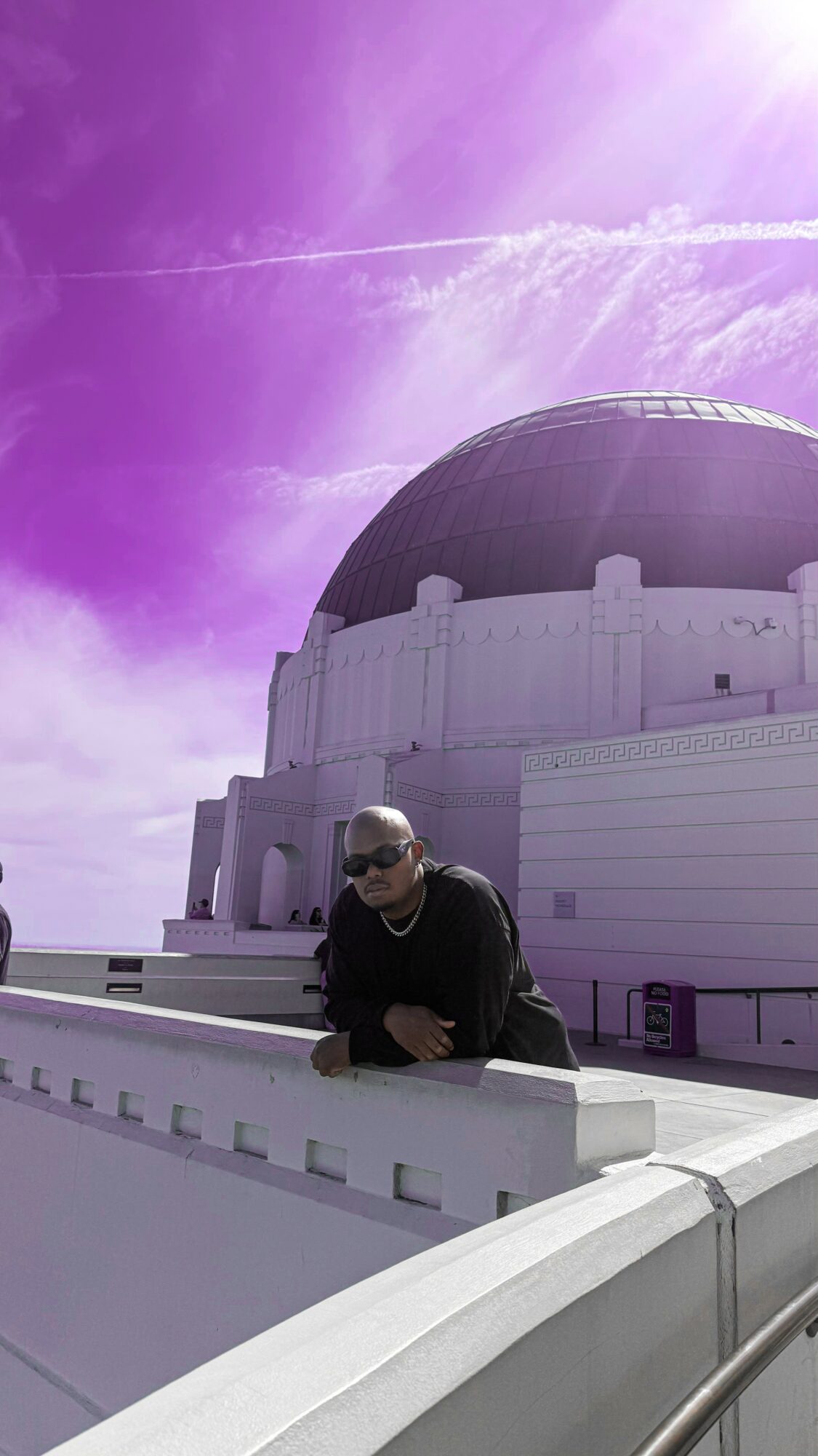 Person wearing sunglasses leaning on a white railing in front of a large observatory dome, with a purple sky and clouds above.