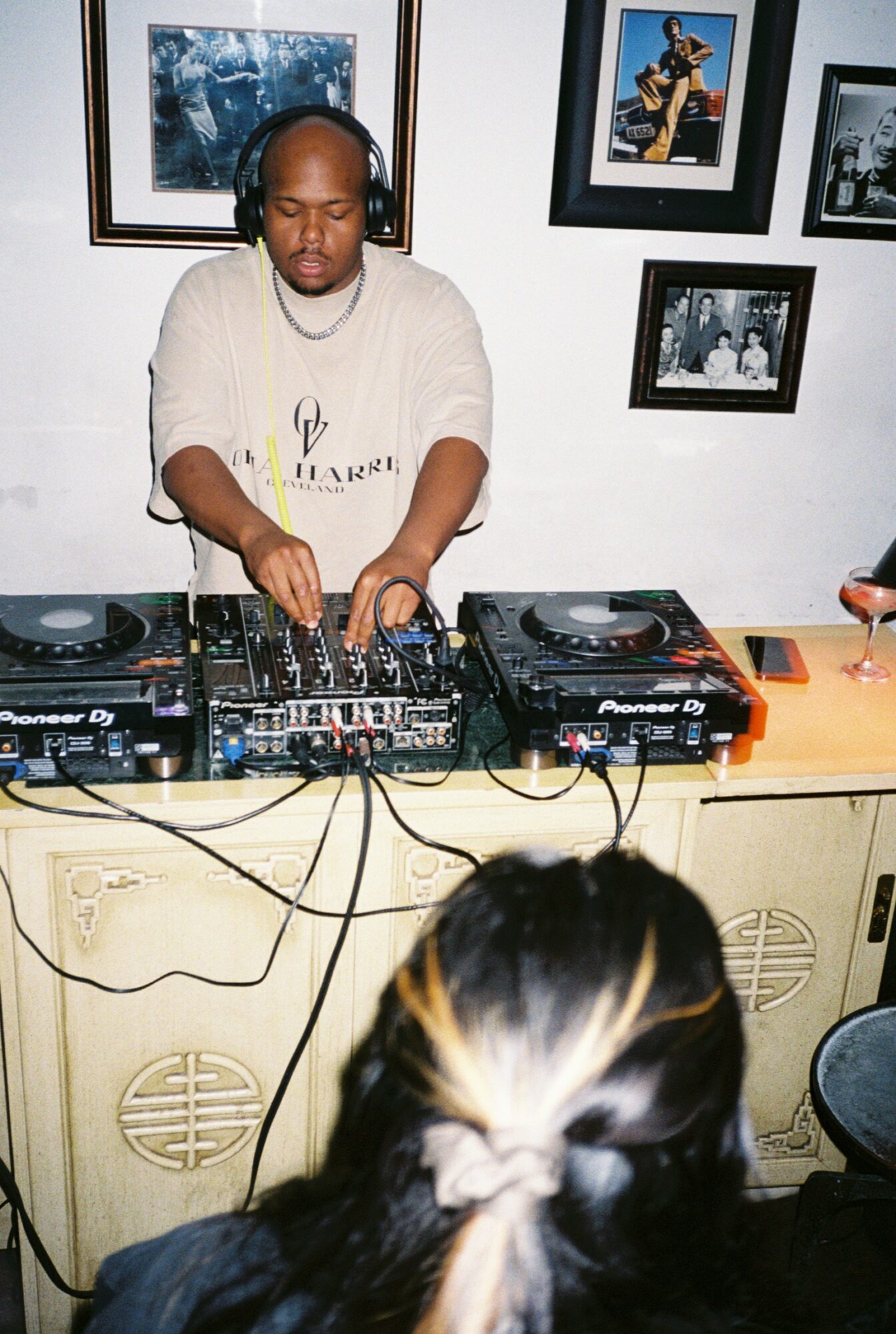DJ wearing headphones operates turntables and mixer in a room with framed photos on the wall, audience in foreground.