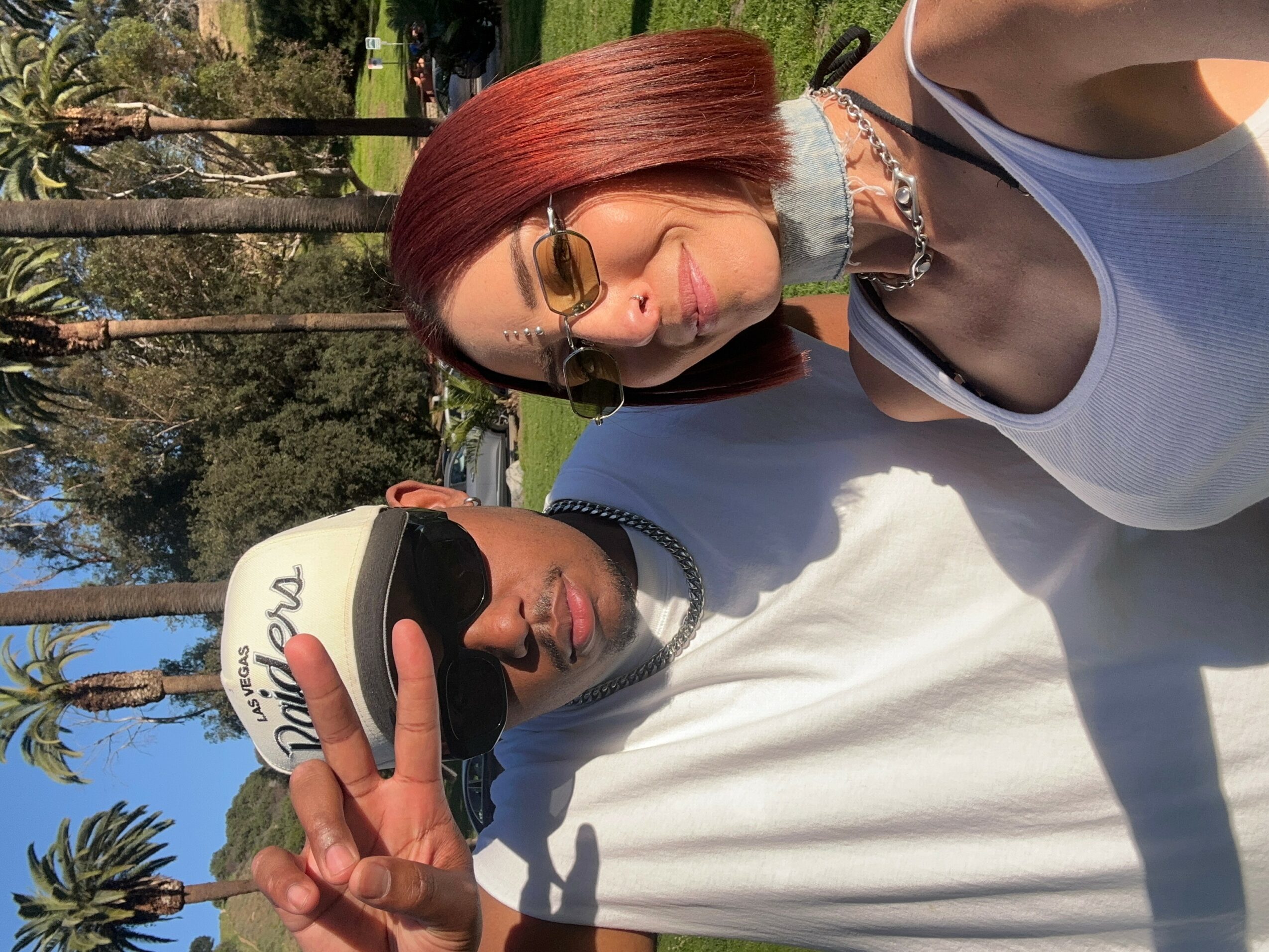 Two people taking a selfie outdoors, one making a peace sign, with trees and blue sky in the background.