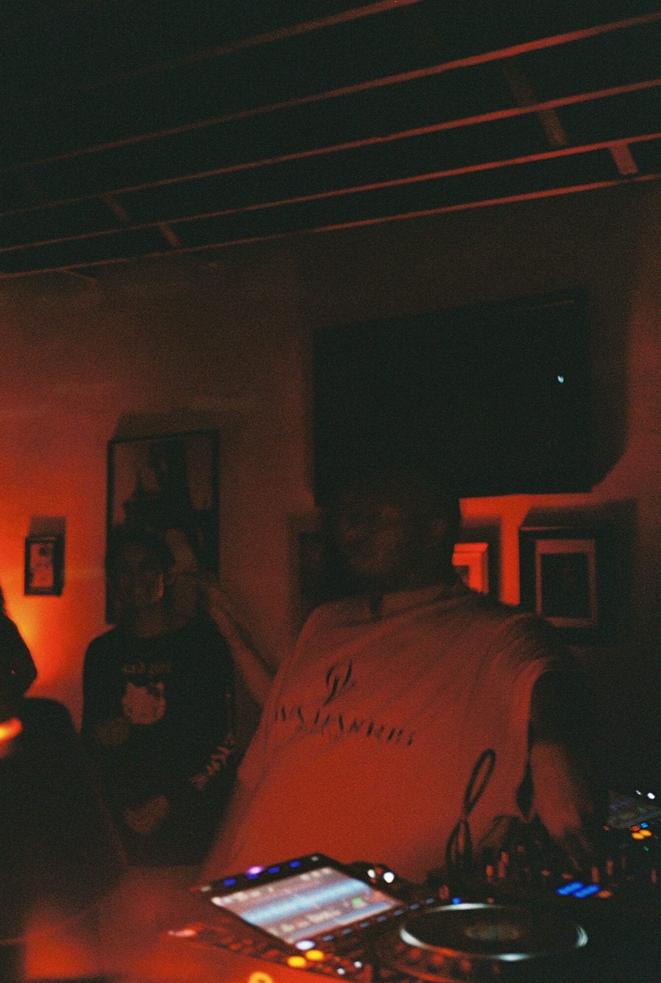 People in a dimly lit room with red lighting, some sitting and some standing, with framed pictures on the wall.
