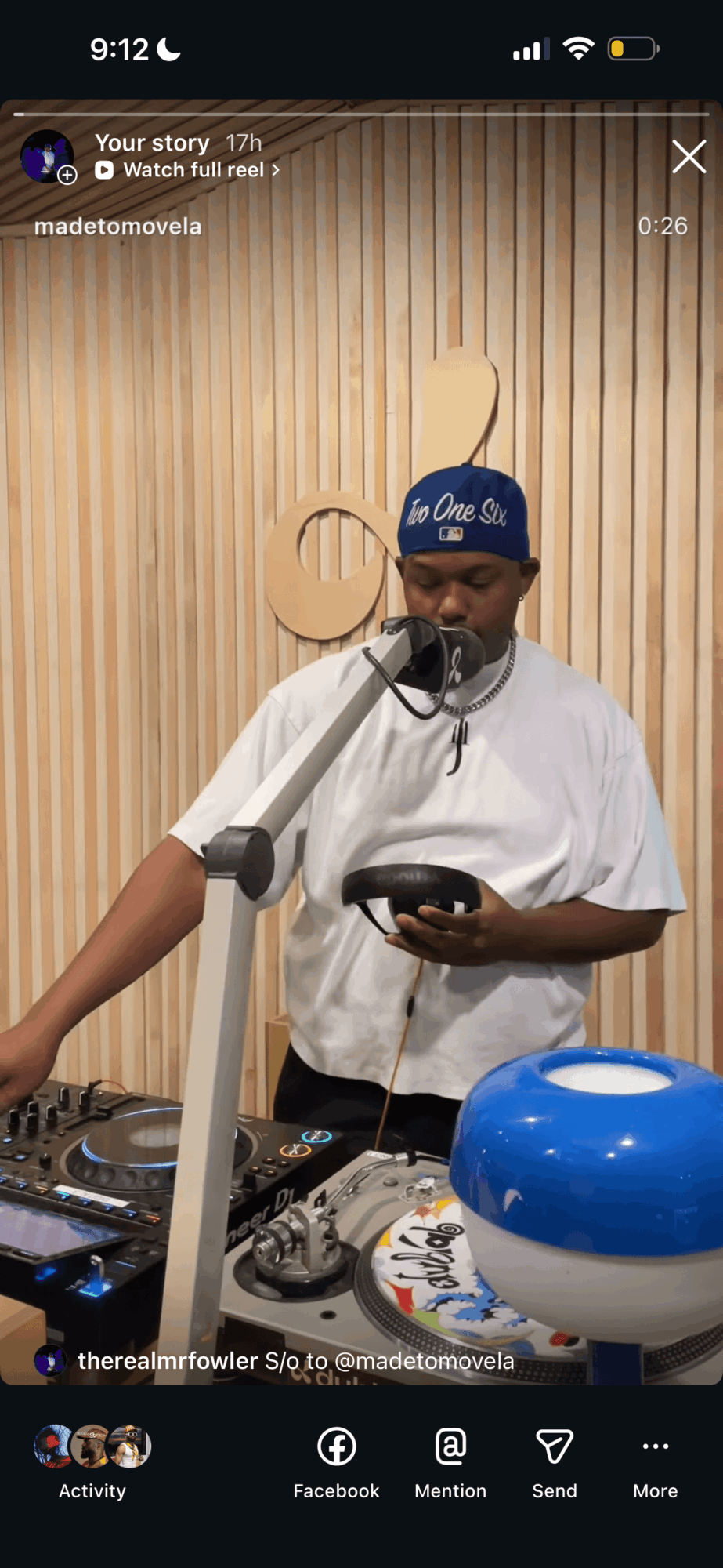 Person wearing a blue cap and white shirt speaking into a microphone, holding a phone, with audio equipment and a blue speaker in front, wooden background.