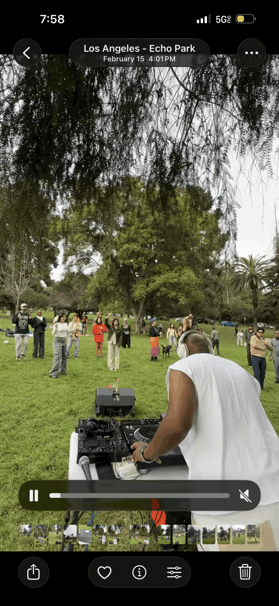 People gather in a park with trees and grass, a person in white is operating a camera, others stand around.