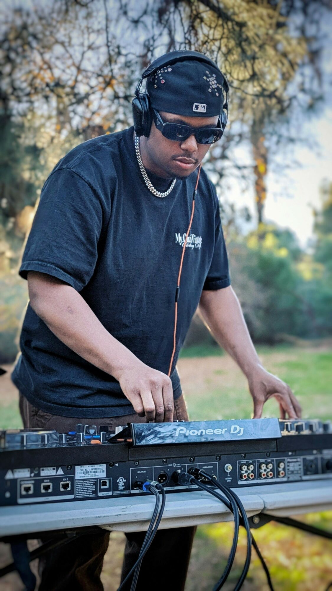Person wearing headphones and sunglasses, operating DJ equipment outdoors with trees and sky in background.