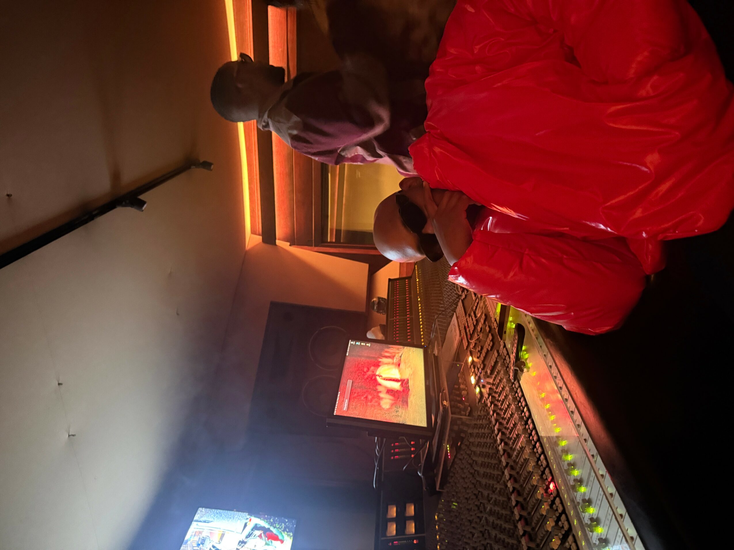 Two people in a dimly lit room with orange lighting, one wearing a red jacket, looking at a screen, with equipment nearby.