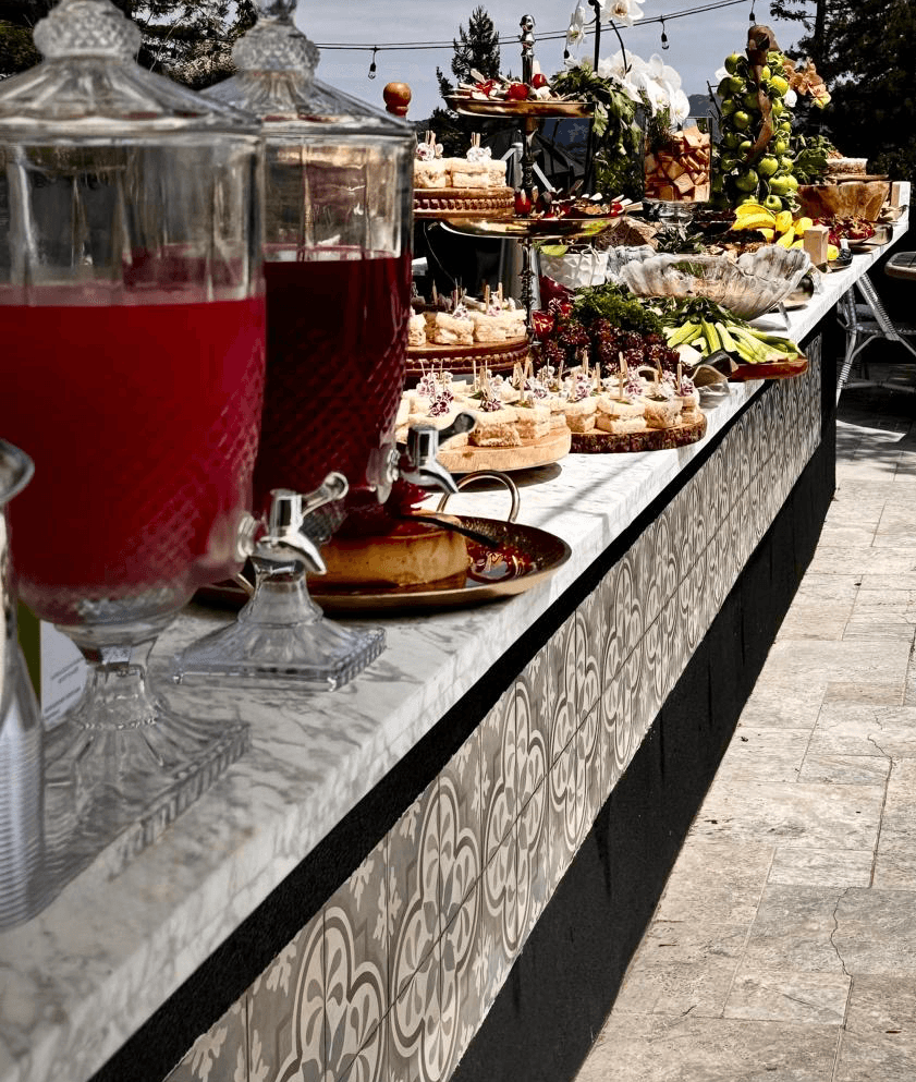 Outdoor buffet table with drinks, desserts, and floral arrangements, set on a patterned black and white tablecloth.