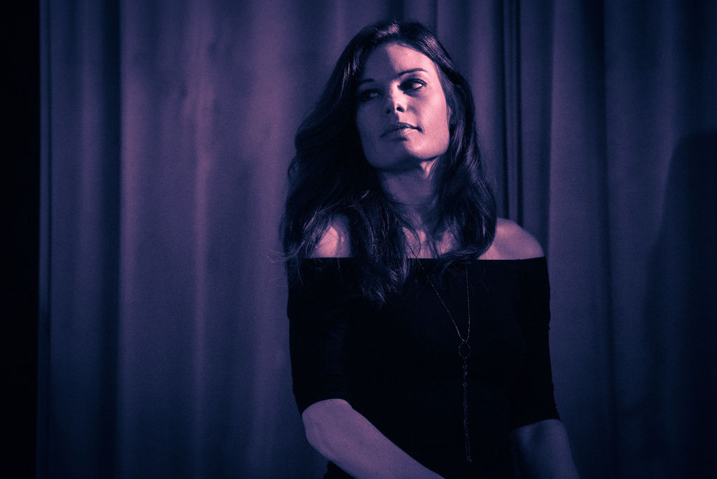 Woman with dark hair and an off-the-shoulder black top stands in front of a dark curtain, looking to the side.