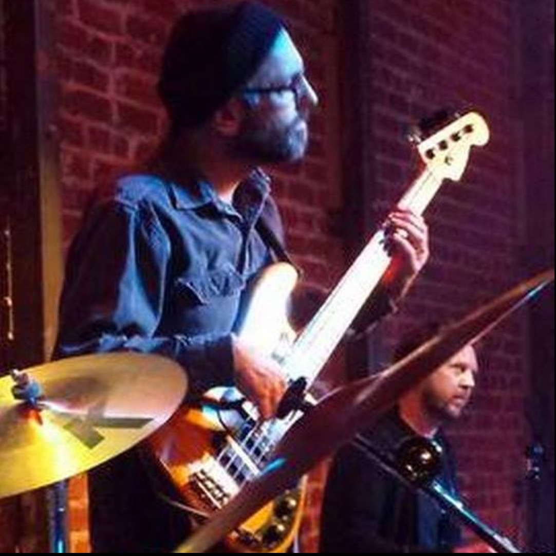 A man with glasses, a beanie, and a beard plays a bass guitar on stage with a brick wall background.