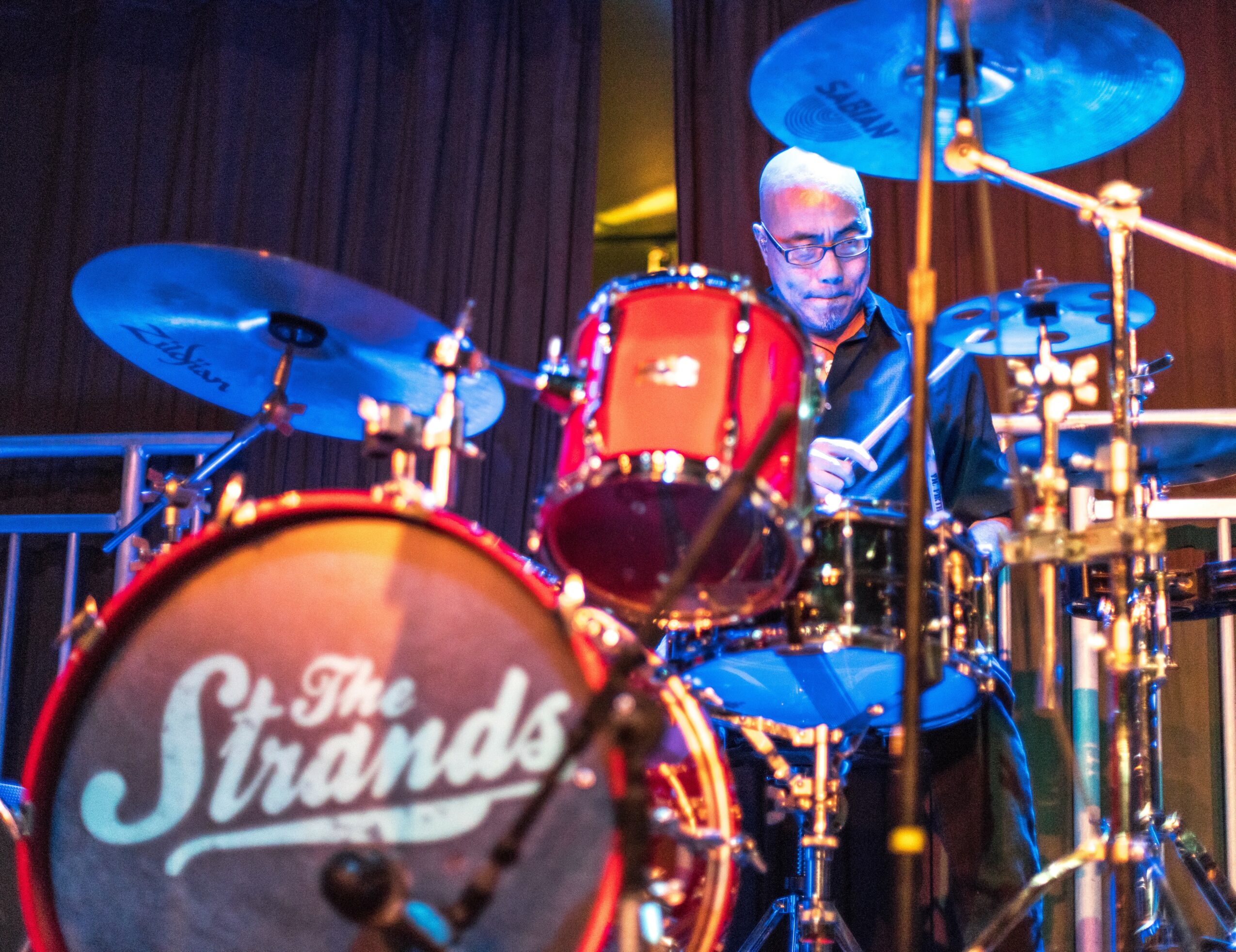 A drummer playing a red drum set on stage with a logo reading 'The Strands' on the bass drum, blue cymbals, and stage lighting.