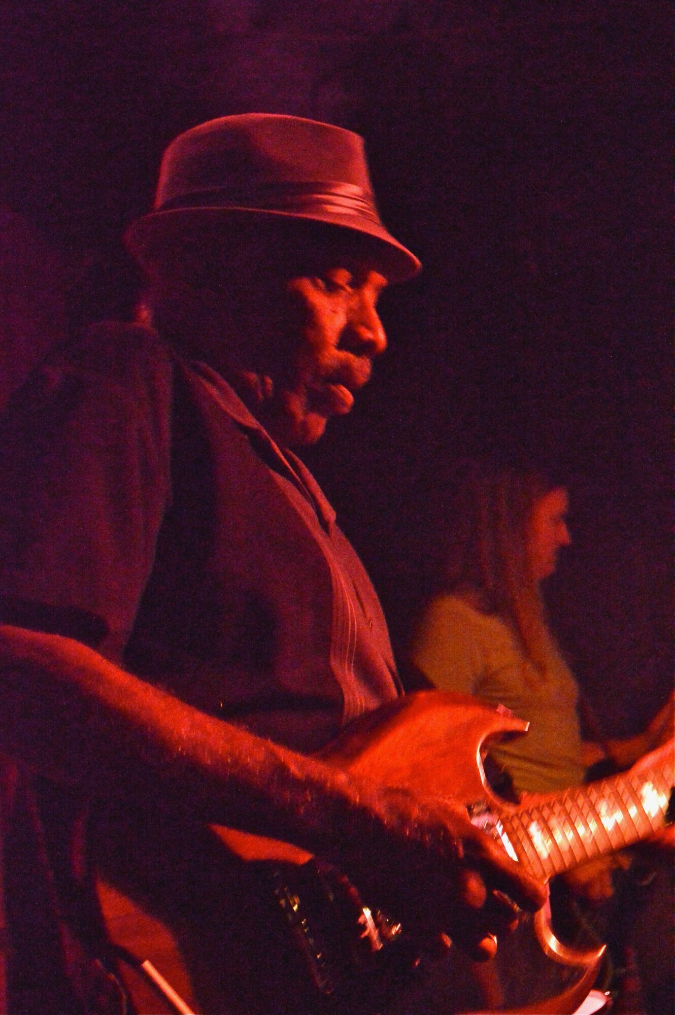 Man wearing a hat playing an electric guitar on stage with a woman in the background, dim lighting.