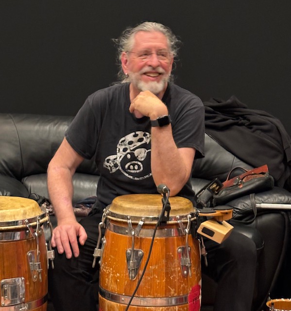 Smiling man with gray hair and beard playing conga drums, sitting on a black couch, wearing a black T-shirt with a skull design.
