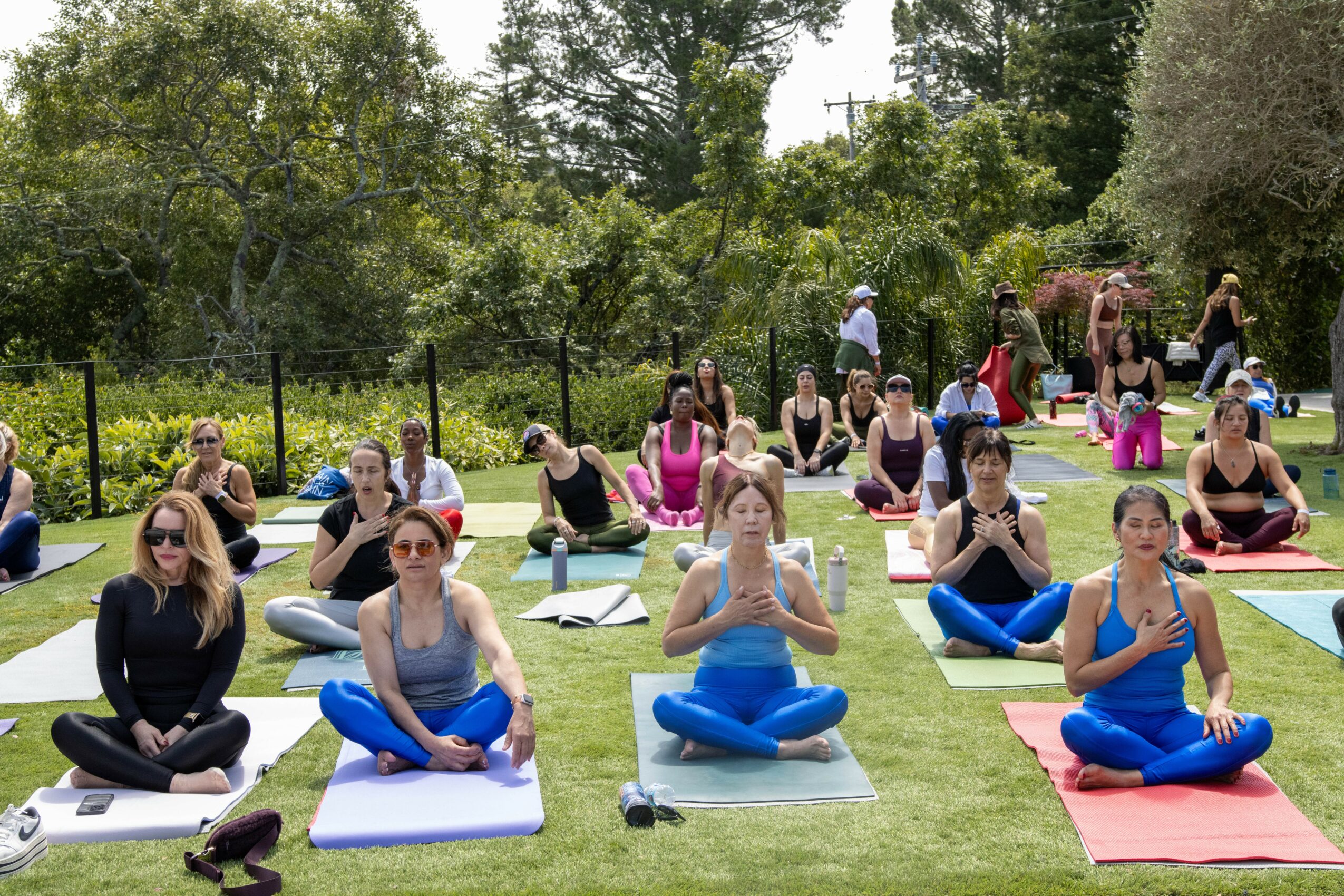 Group of people practicing yoga outdoors on mats, sitting cross-legged with hands in prayer position, surrounded by trees and greenery.