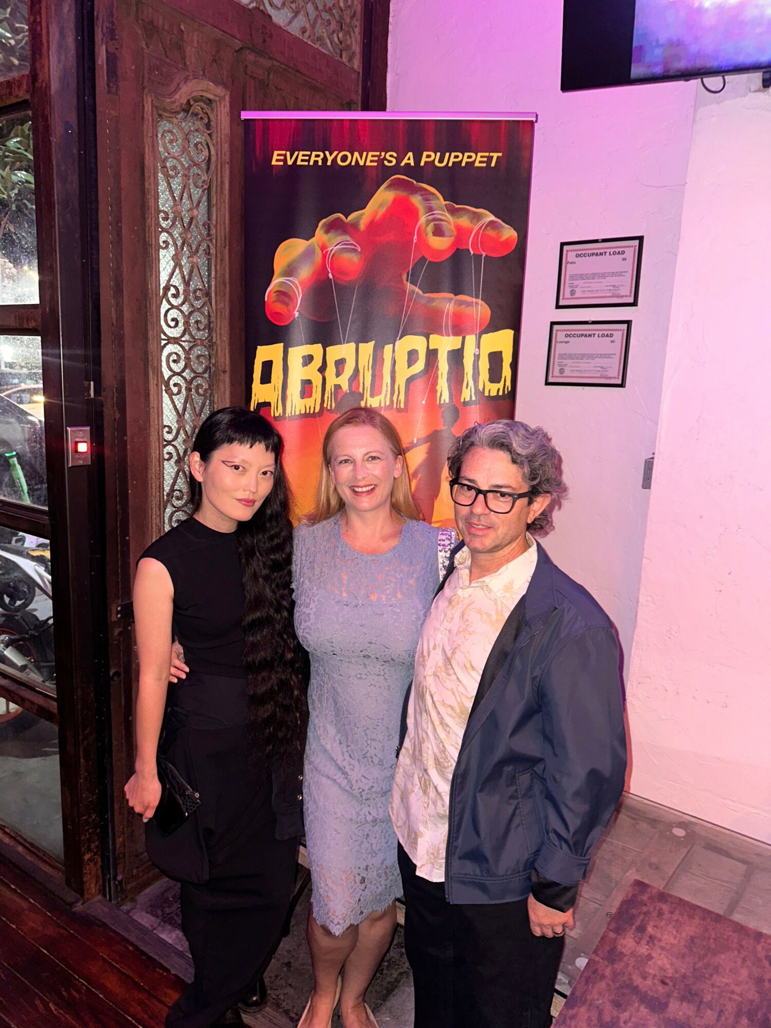 Three people standing indoors in front of a poster for the movie 'Brazil' with a fiery background, two women and one man, all smiling.