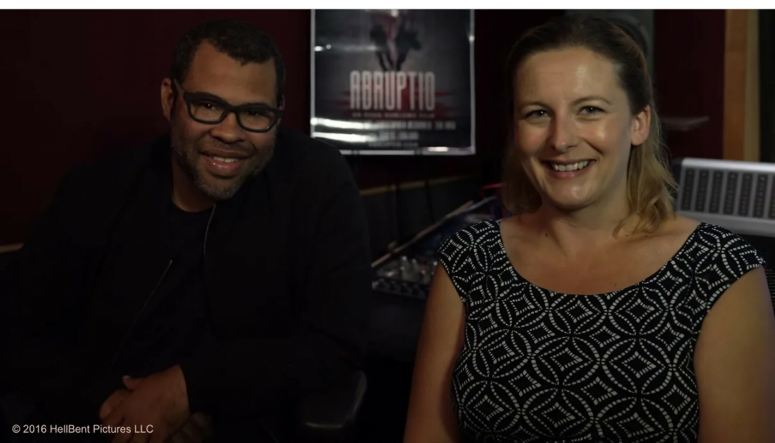 Two people sitting indoors, smiling, with a movie poster in the background.