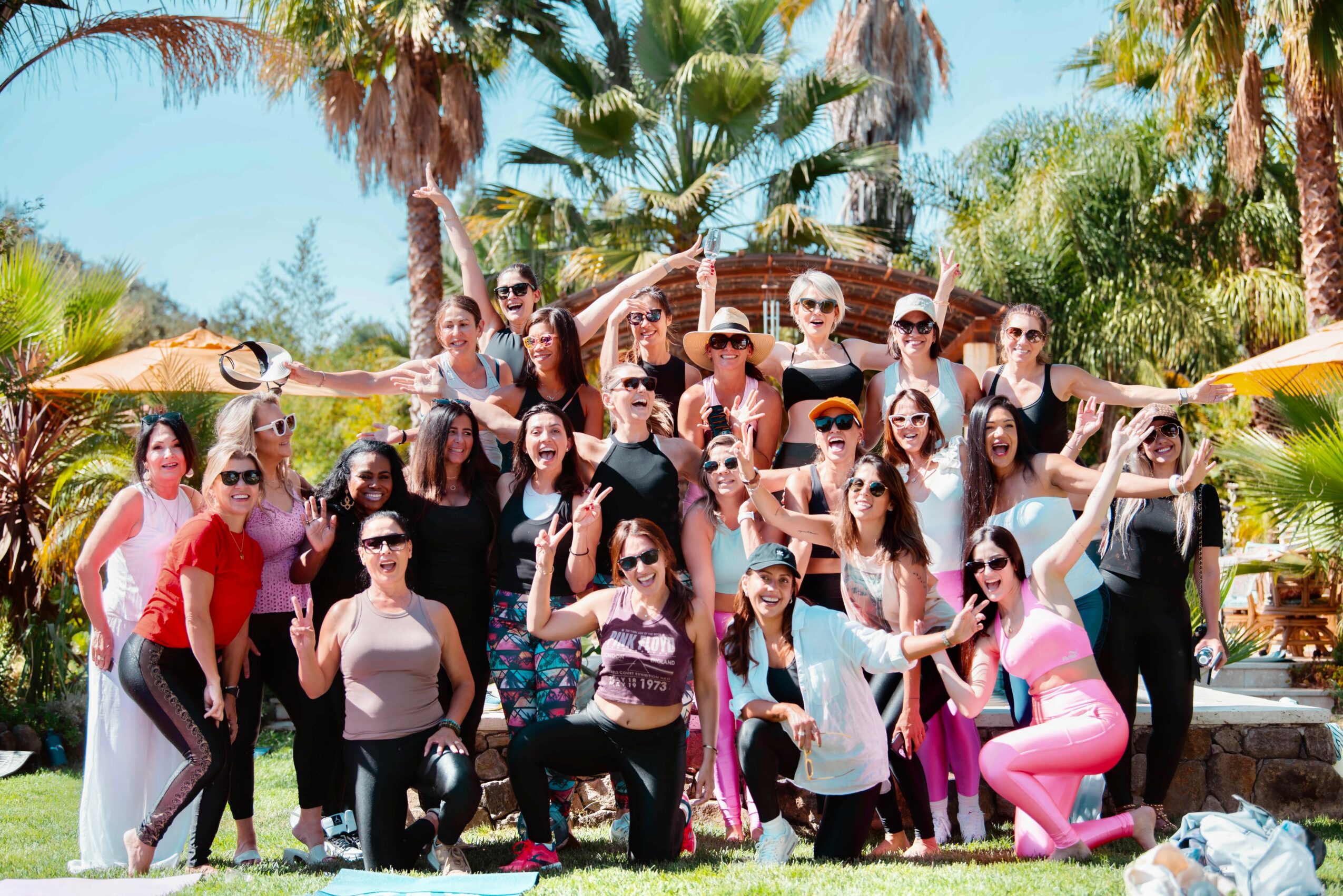 Group of people outdoors with palm trees, some holding umbrellas, smiling and posing for a photo.