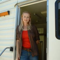 Woman standing in doorway of a camper, smiling, wearing a red shirt and brown jacket.