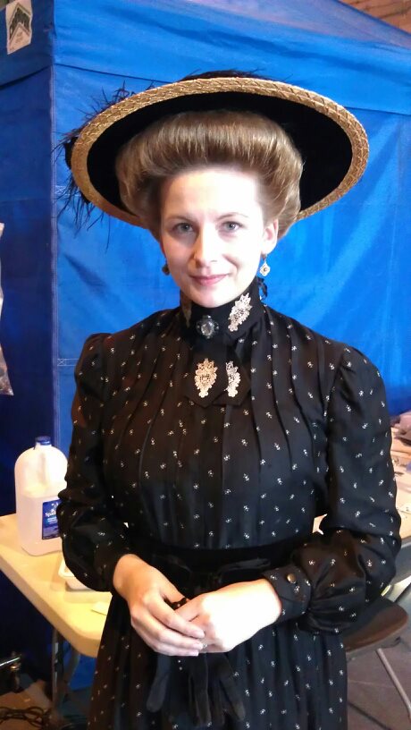 Woman wearing a large hat and vintage-style black dress with polka dots, standing indoors with a blue background.