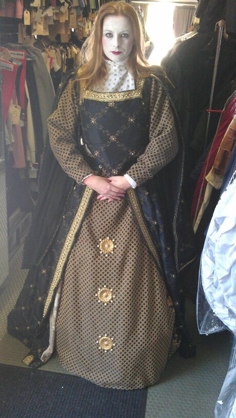Person dressed in historical costume with long patterned skirt, black and gold bodice, and sheer polka-dot sleeves, standing in a clothing store.
