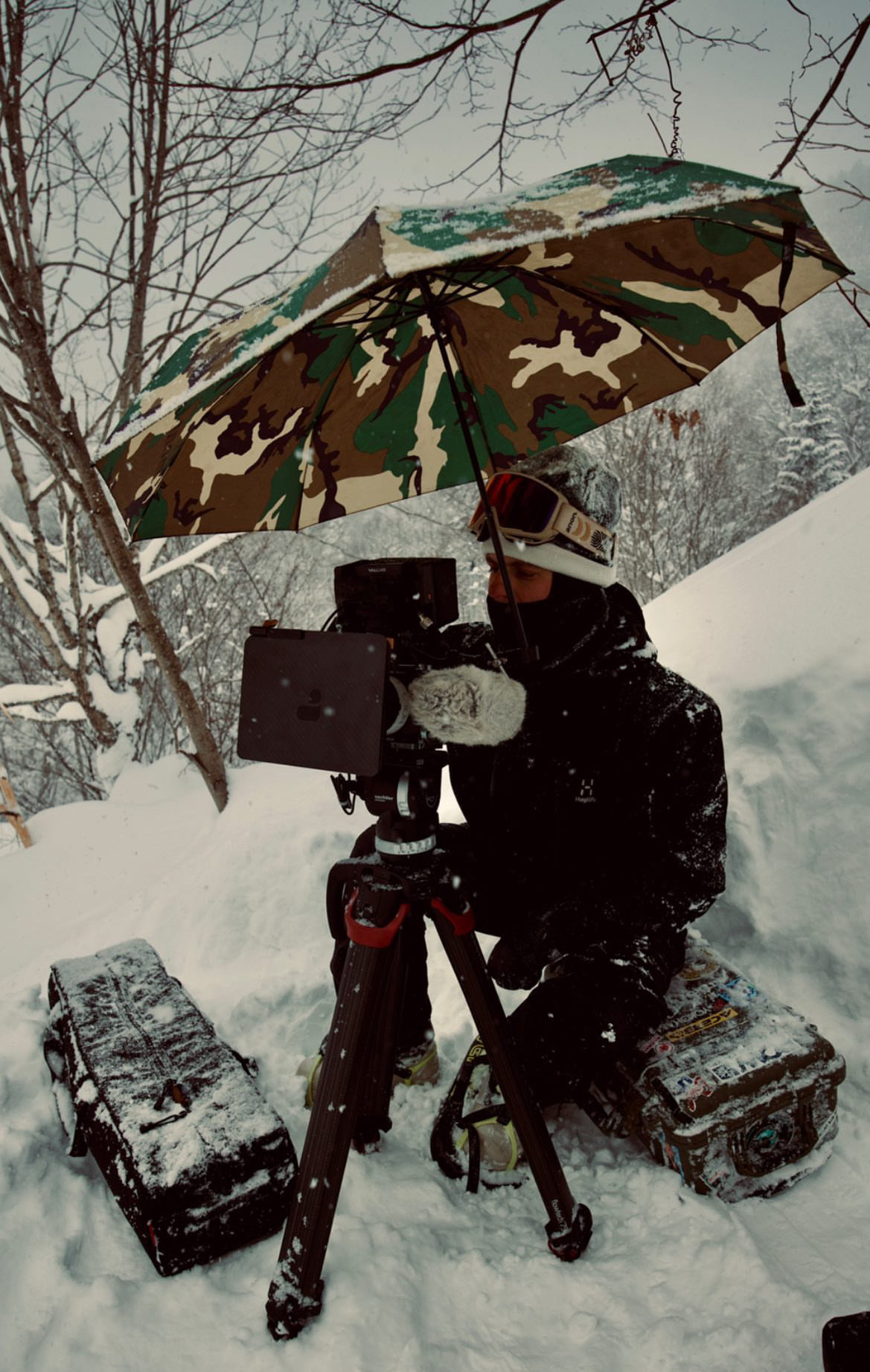 Person operating camera outdoors in snowy environment, holding a camouflage umbrella, with equipment and snow-covered trees around.