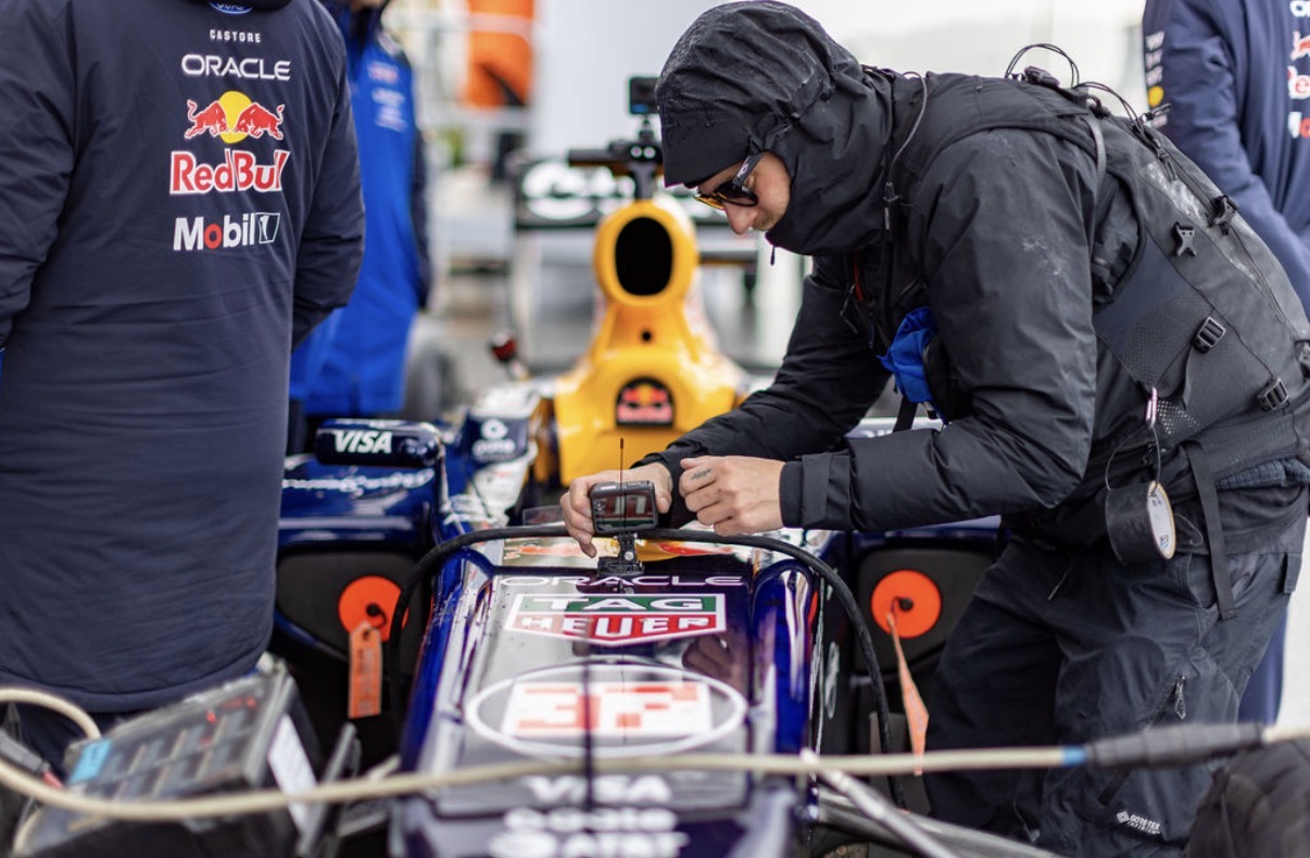 Person in black jacket and hood working on a race car in a garage, with team members and equipment around.
