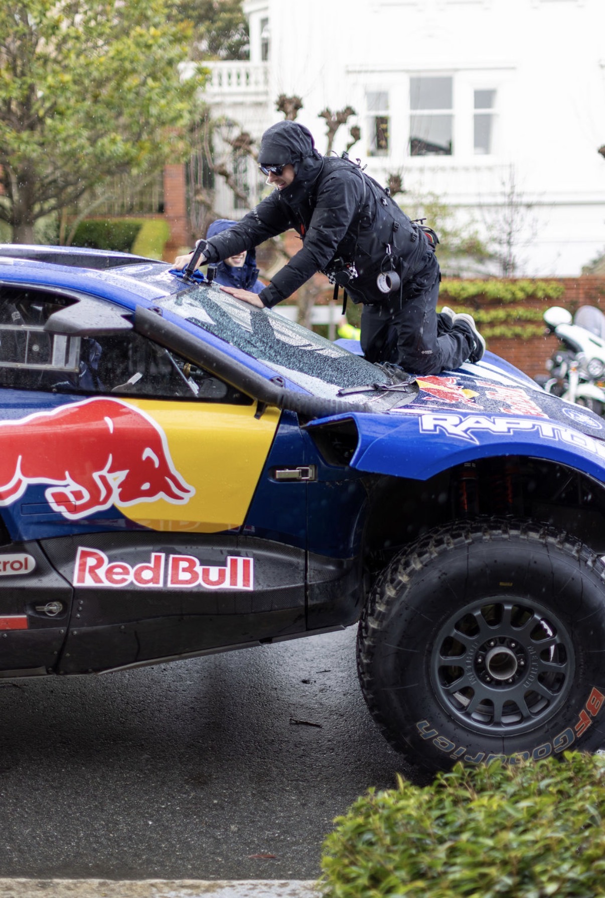 Police officer kneeling on a race car with Red Bull logo, inspecting or working on it, on a street with houses and greenery.