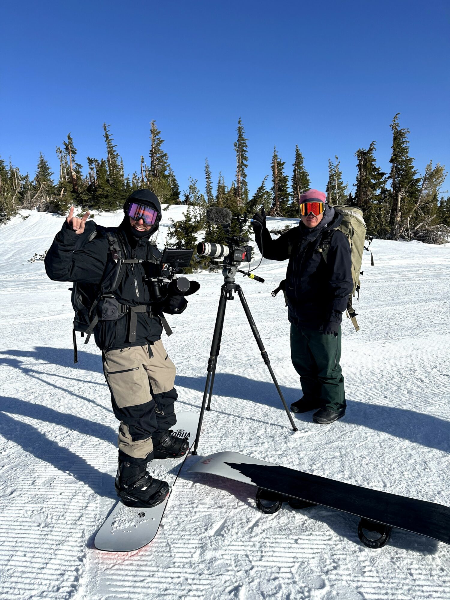 Two people in winter gear stand on snow with a camera on a tripod, trees and blue sky in background.