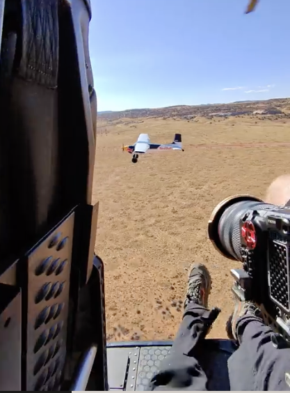 Person sitting in a helicopter looking at a small aircraft on the ground in an open field.