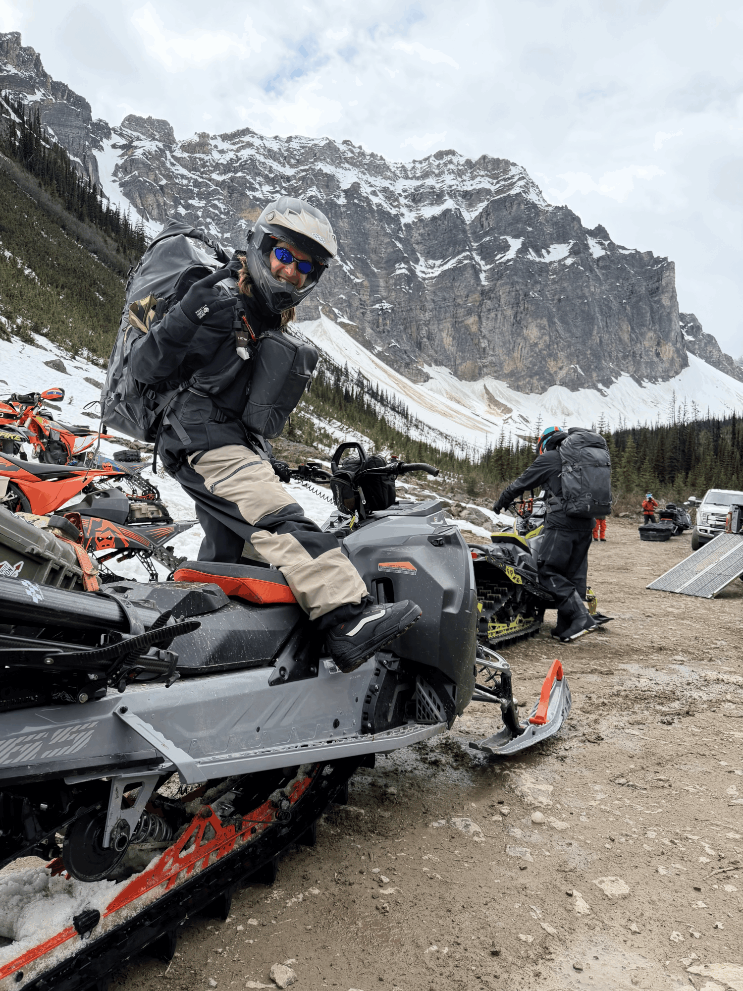 Person in motorcycle gear sitting on a snowmobile in a mountainous area with snow and trees, cloudy sky, other people and vehicles in background.