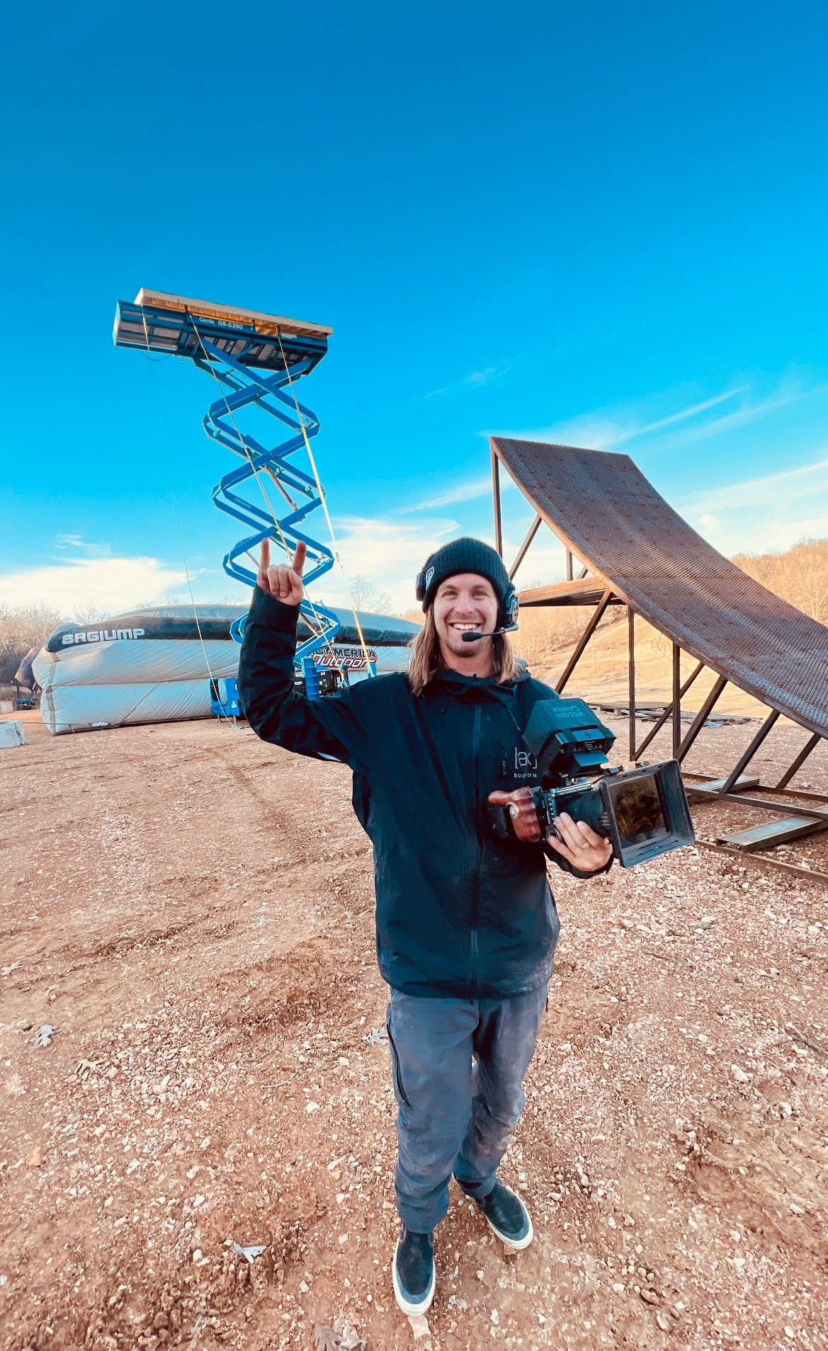 Person smiling and waving outdoors, holding a camera, with a ramp and a scissor lift in the background, under a clear blue sky.