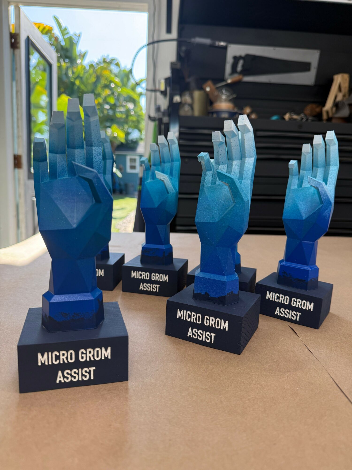 Three blue robotic hand sculptures on black bases labeled 'Micro Groom Assist' displayed indoors with a window and outdoor greenery in background.