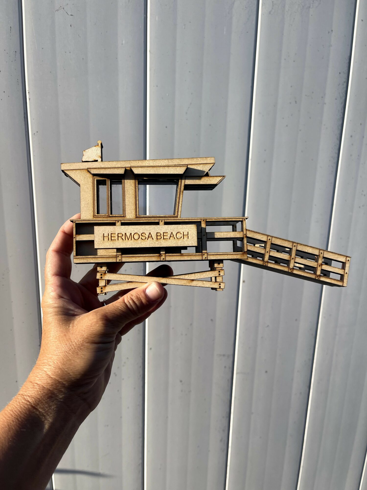 Hand holding a wooden model of a building with a sign reading 'HERMOSA BEACH' against a corrugated metal background.