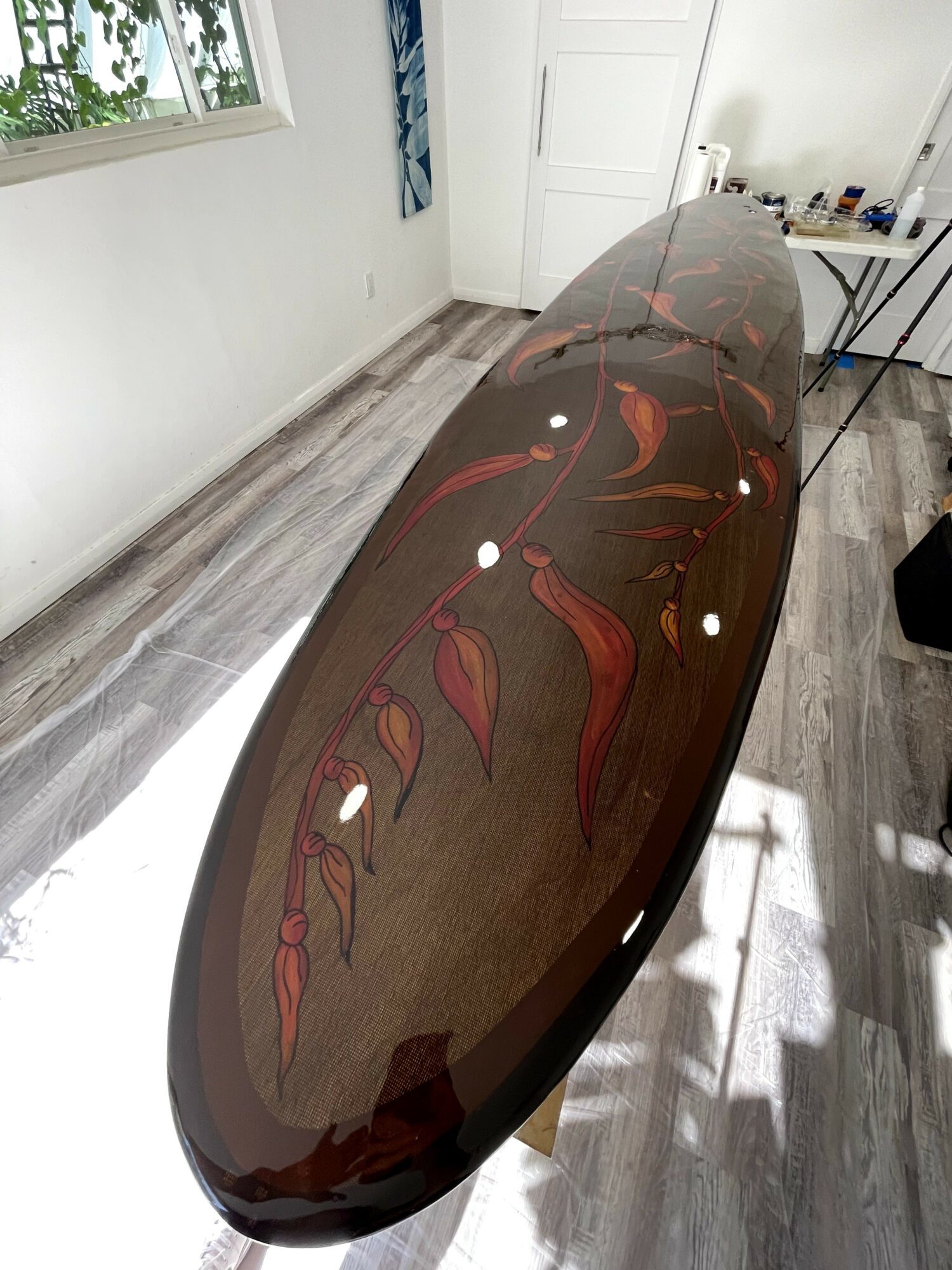 Long, oval wooden surfboard with red and orange leaf-like designs, resting on a wooden floor indoors.