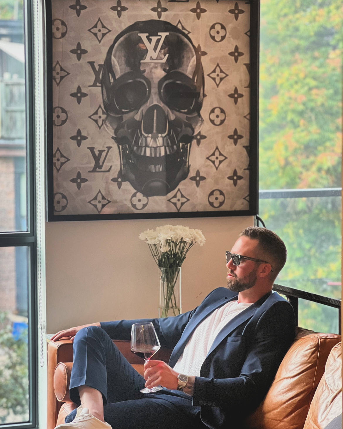 Man in suit with sunglasses sitting on a sofa, holding a glass, with a skull artwork and flowers behind him.