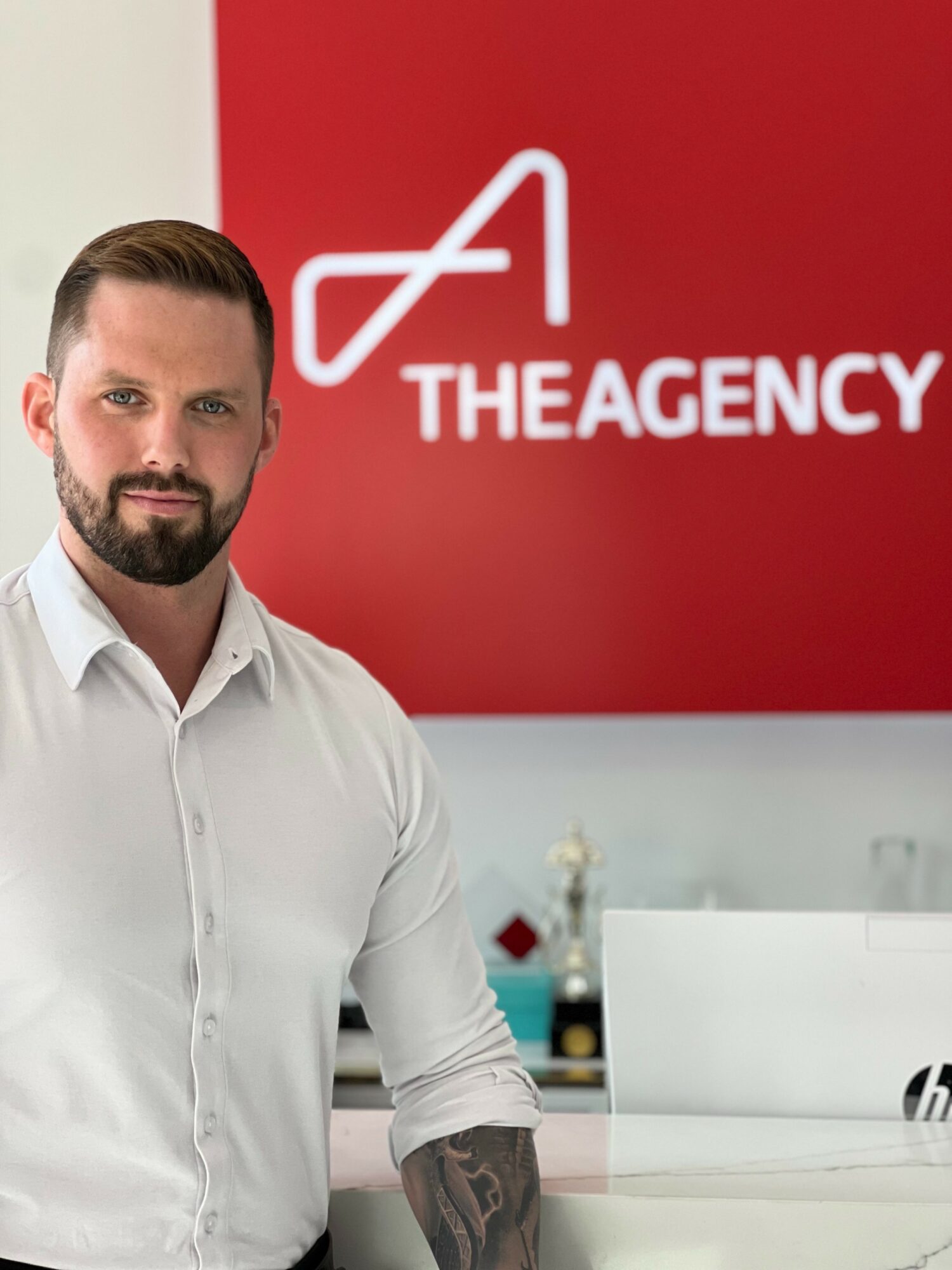 Man with beard and white shirt standing in office with red wall and logo reading THE AGENCY.