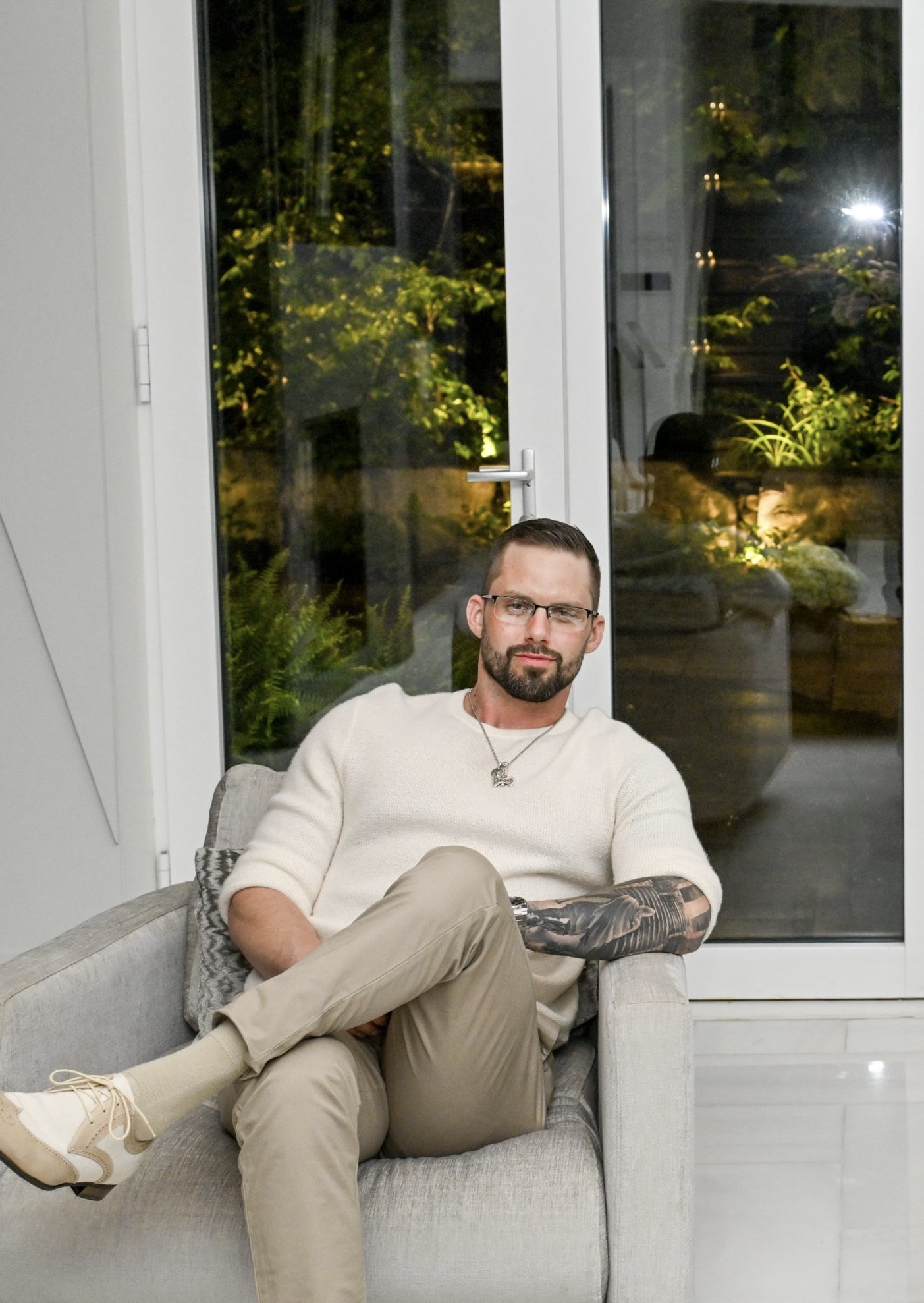 Man with glasses and beard sitting on a sofa in a room with large glass doors showing a night view outside.