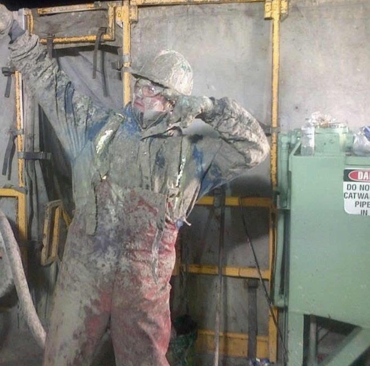 Worker covered in dust or paint, wearing safety gear, standing near industrial equipment and yellow safety rails.
