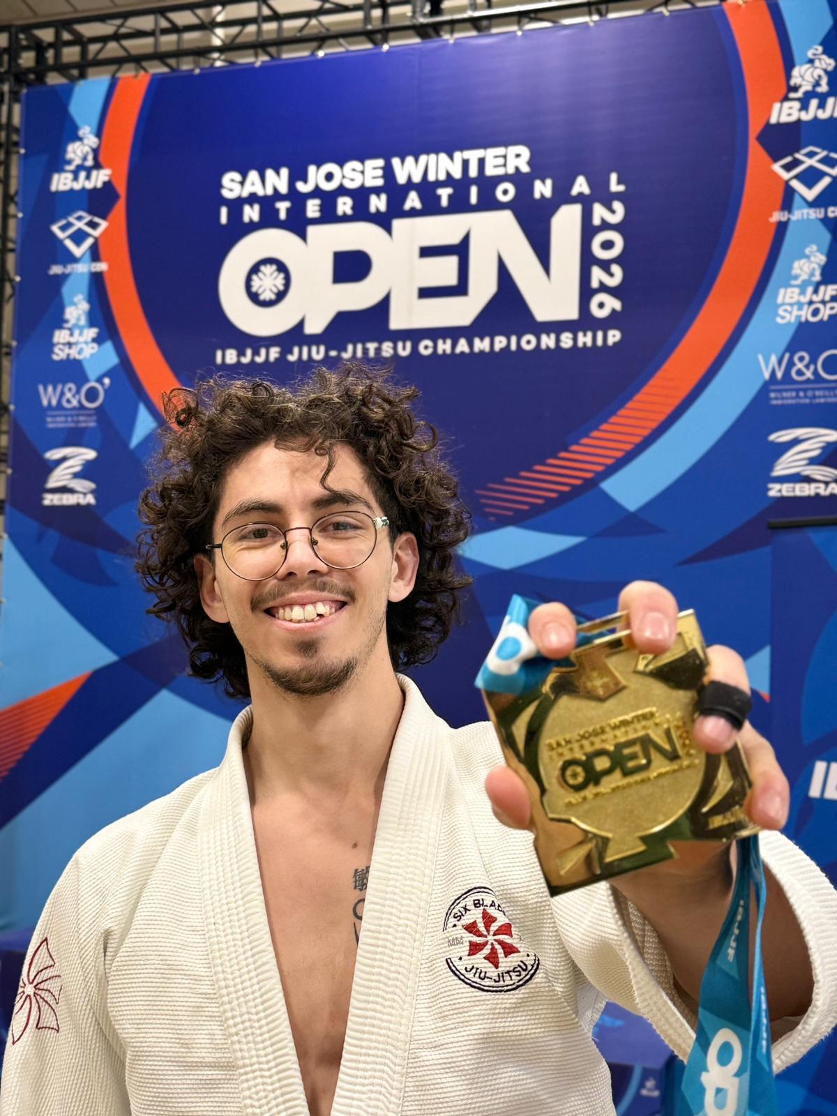 Person with curly hair smiling, holding a gold medal, in front of a blue banner for San Jose Winter Open 2022, jiu-jitsu championship.