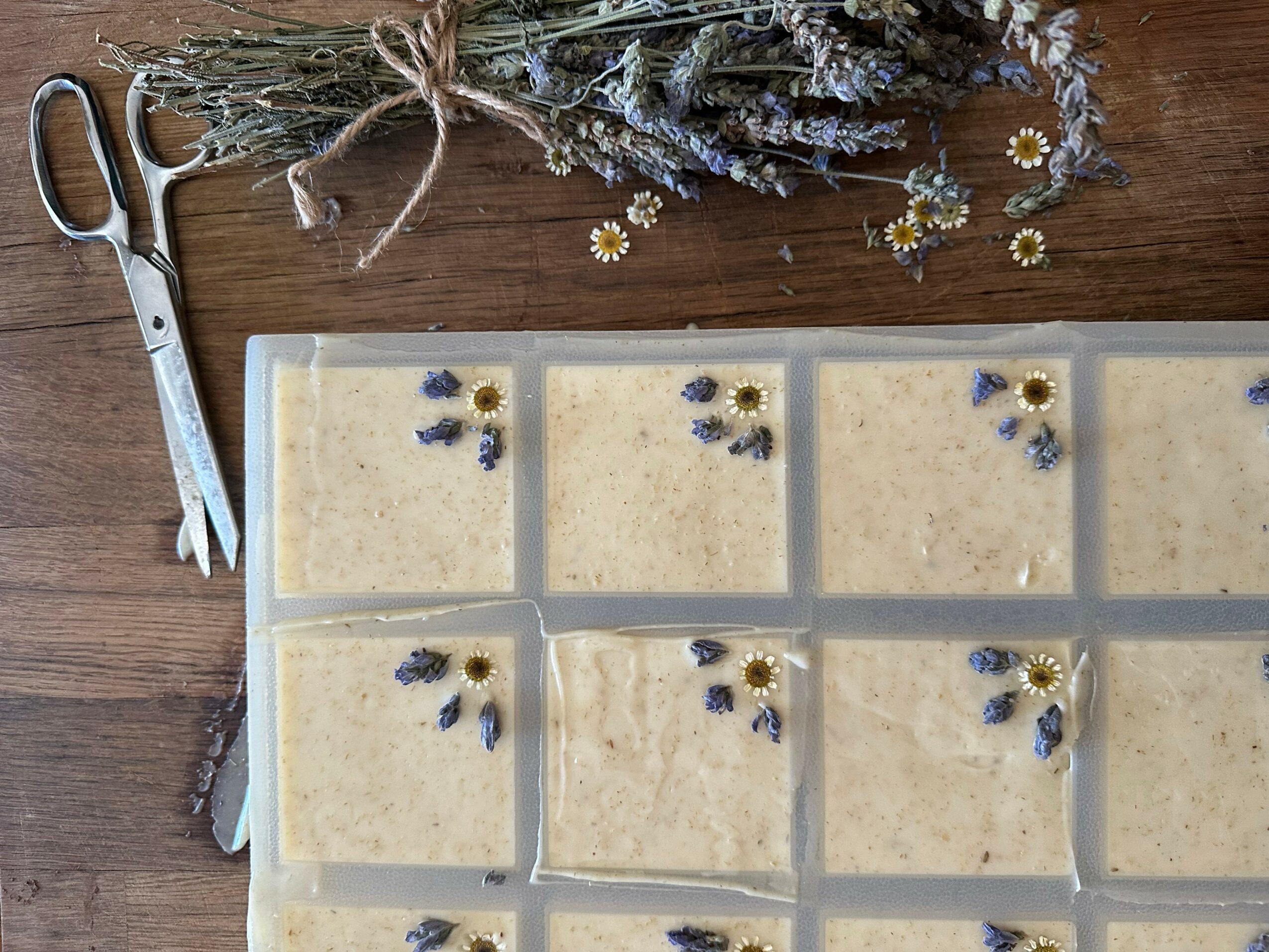 Unbaked soap in individual molds decorated with dried flowers and herbs, on a wooden surface with scissors and dried flowers nearby.