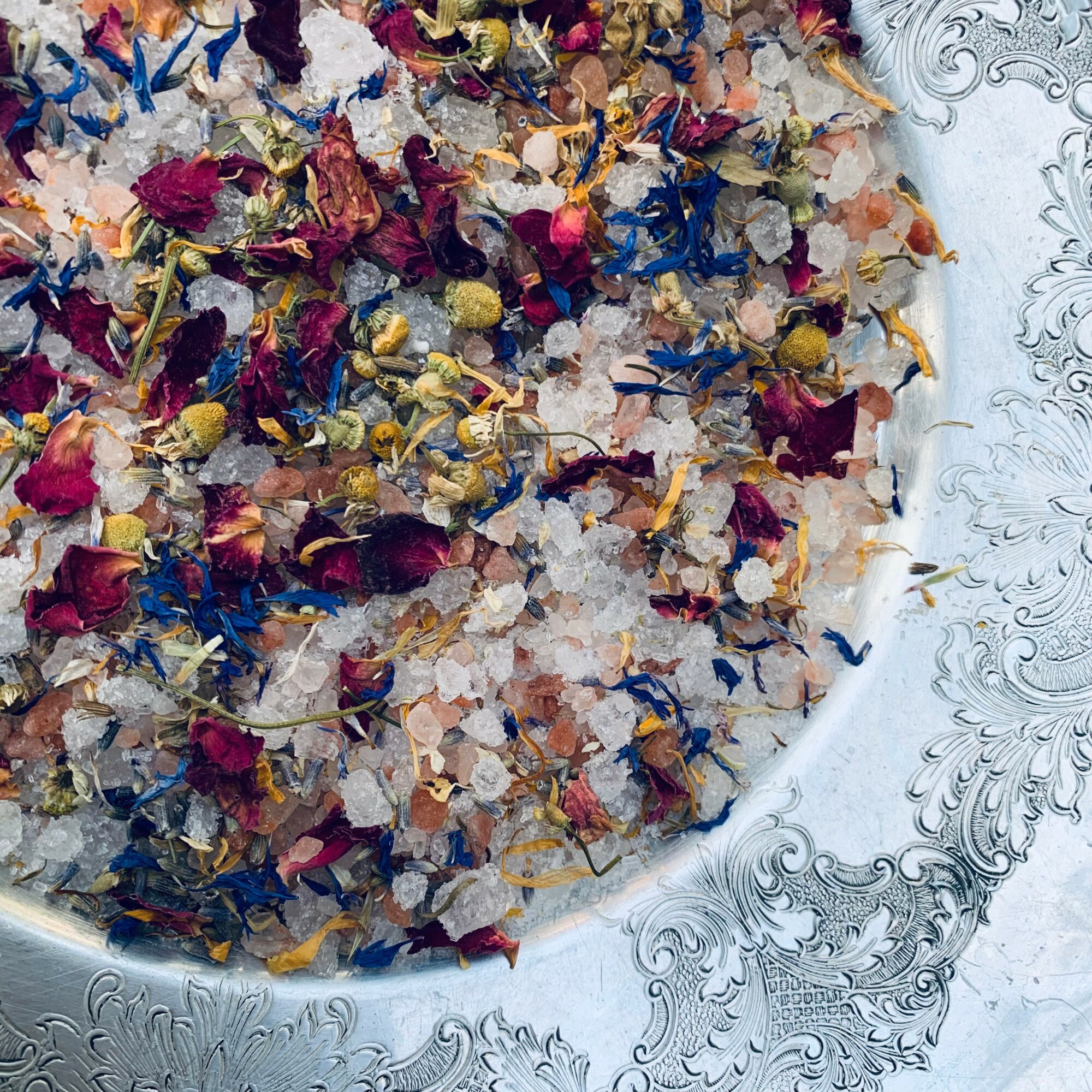 Close-up of a decorative silver plate with colorful dried flower petals and salt crystals, ornate border design.
