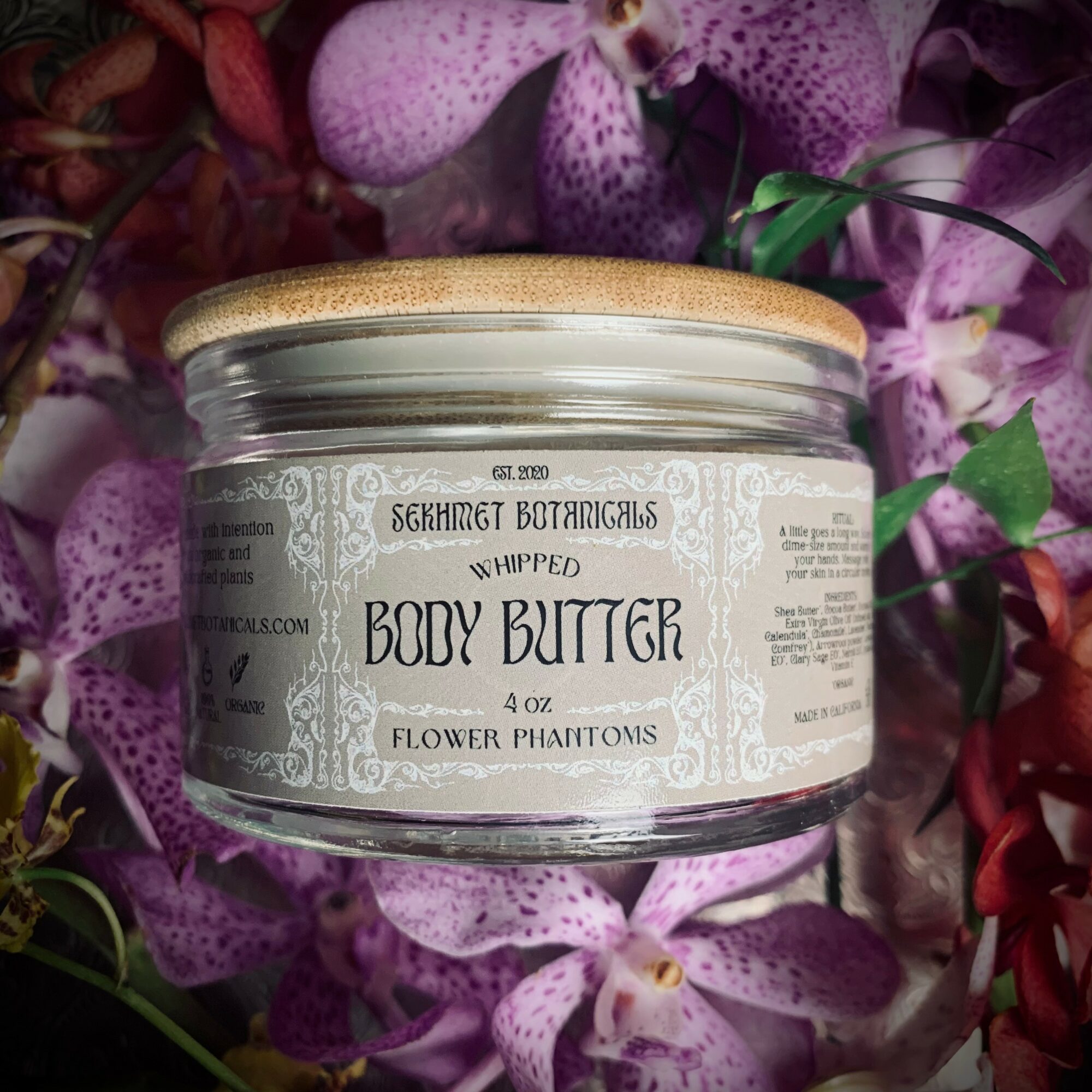 A glass jar of body butter with a gold lid, surrounded by purple and pink flowers, label reads 'Whipped Body Butter'.