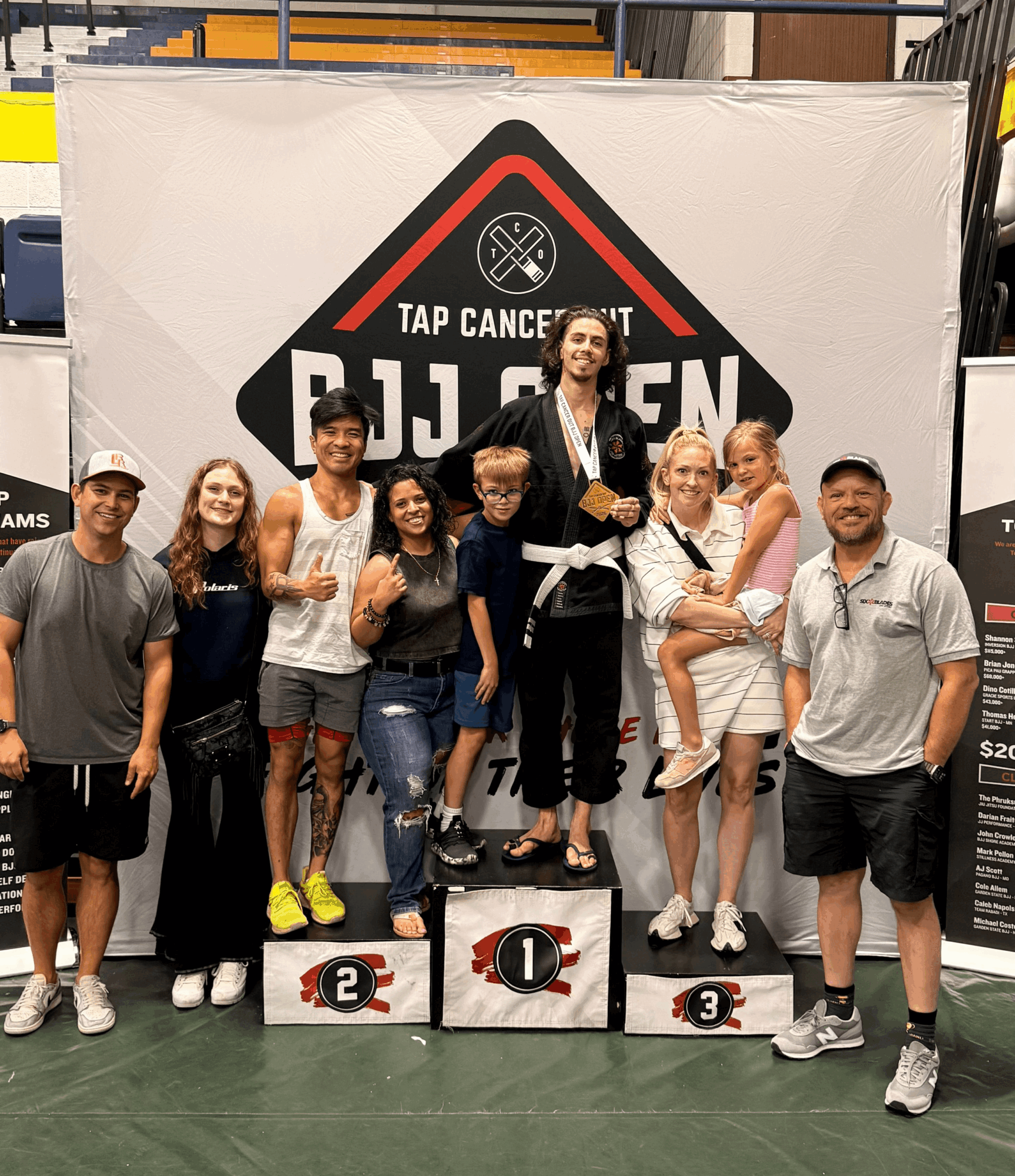 Group of people standing on a winners' podium with a woman holding a child, celebrating at a tap dance event, with a large banner in the background.