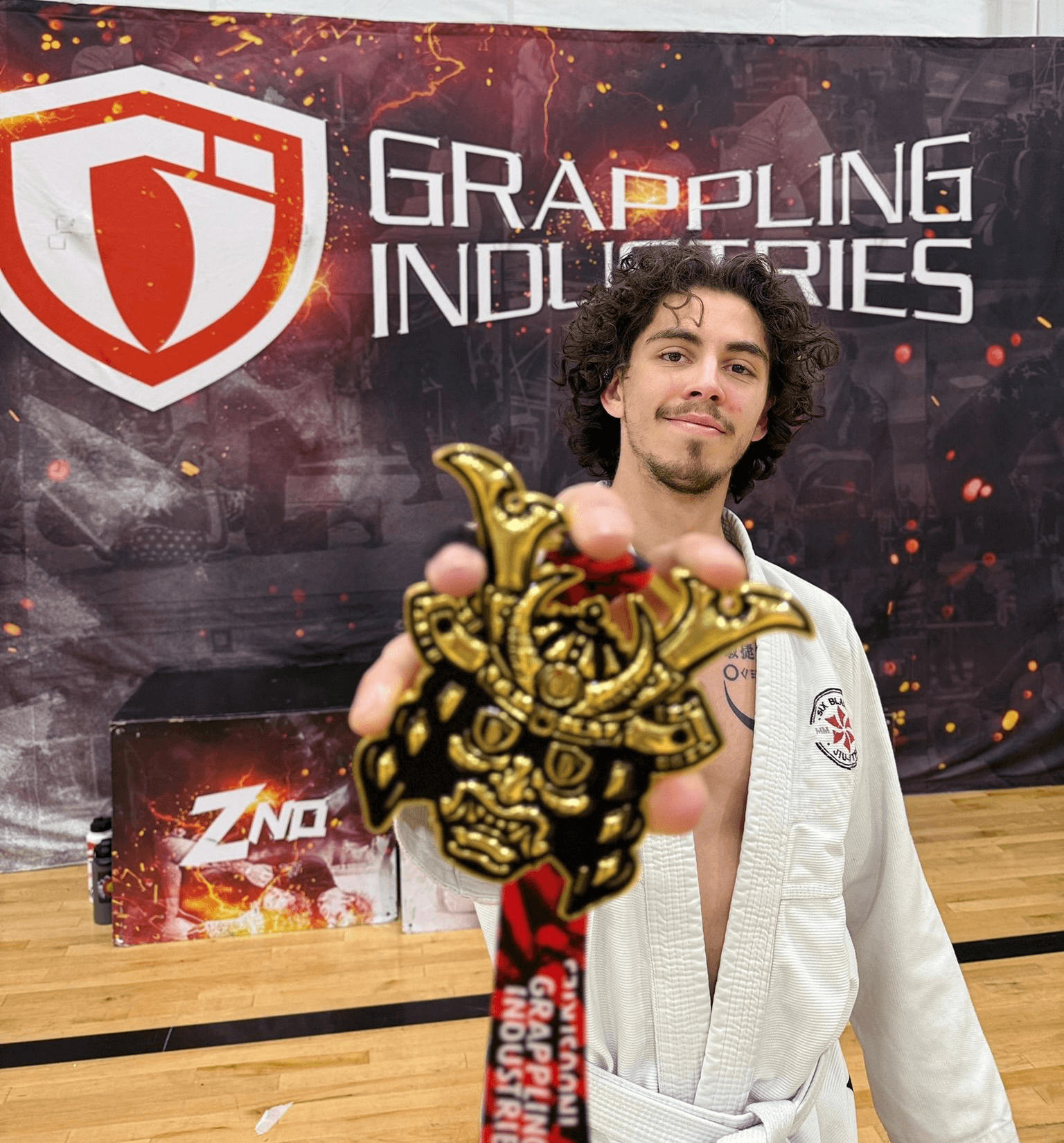 Young man with curly hair in a white martial arts gi, holding a gold medal, standing in front of a grappling industries banner.