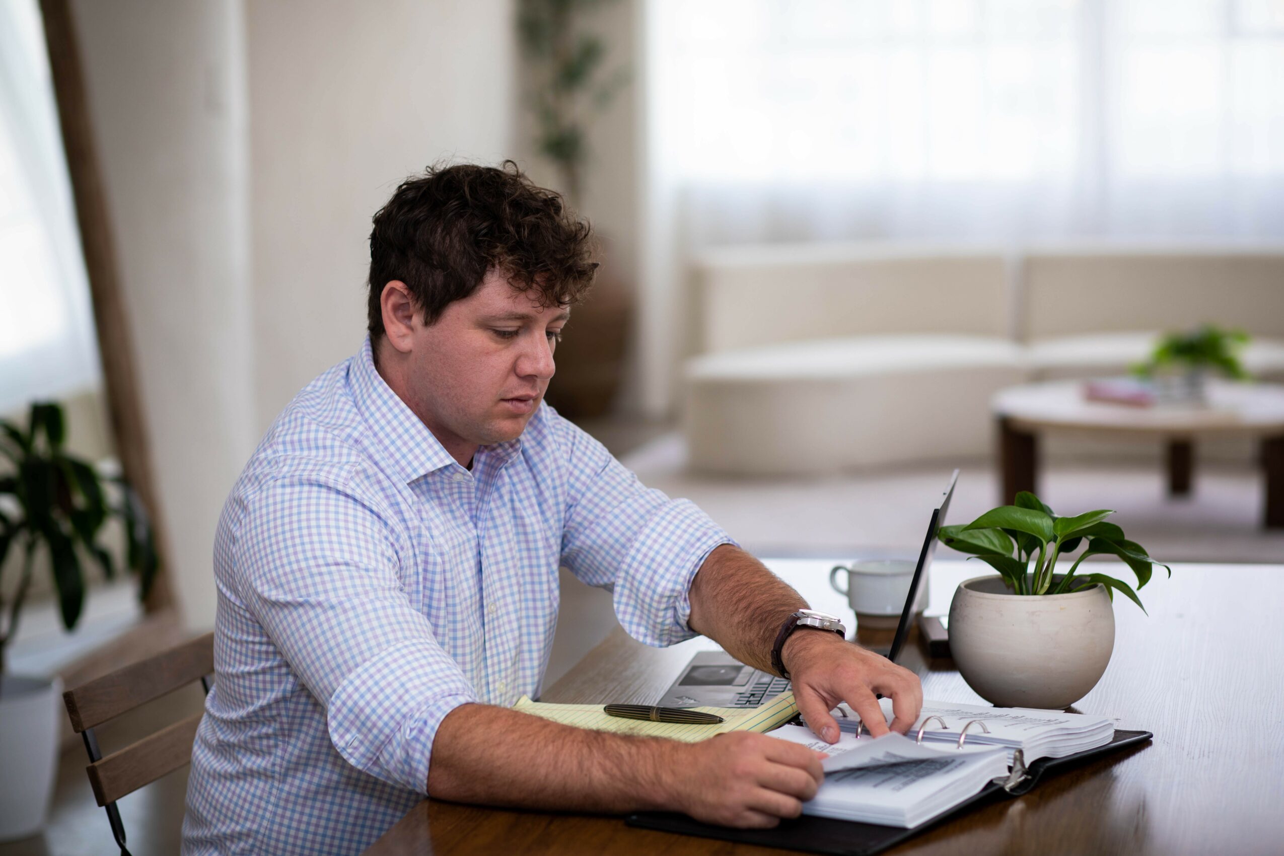 Young man with dark curly hair reading a book at a table in a bright room, with a potted plant nearby.