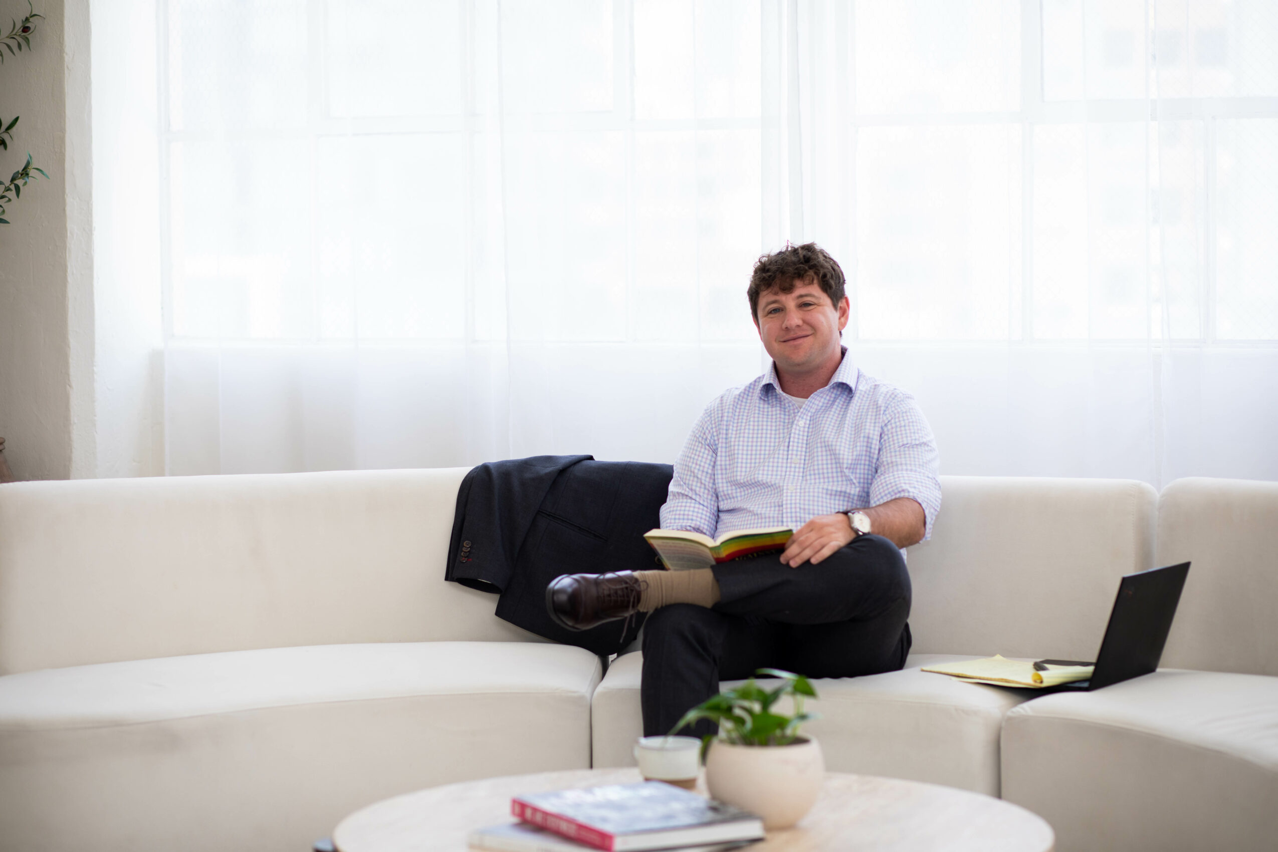 Man sitting on a white sofa holding a book, with a laptop and a plant nearby, in a bright room.