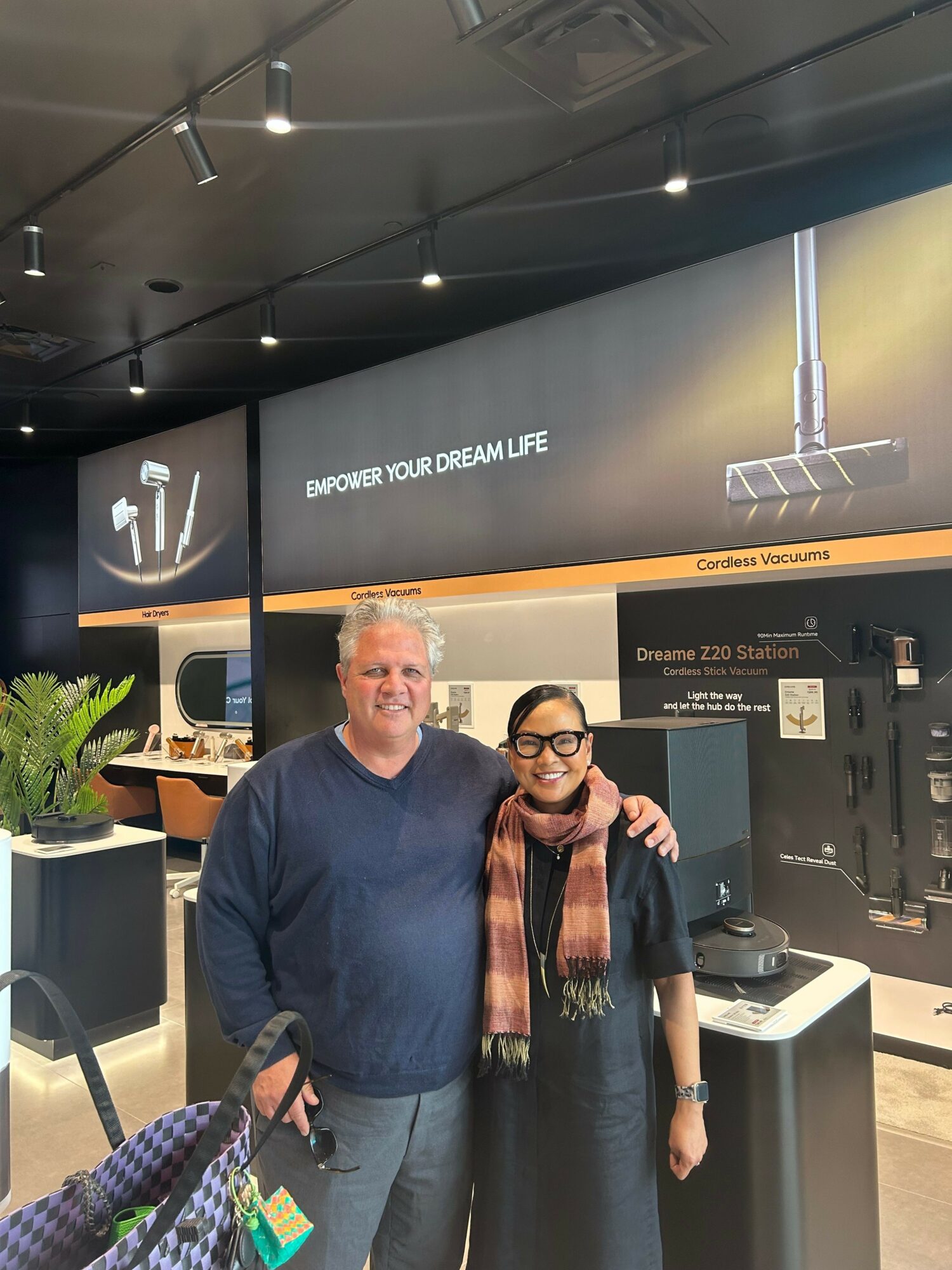 Two people standing in front of a coffee machine at a cafe, smiling, with a digital menu behind them.
